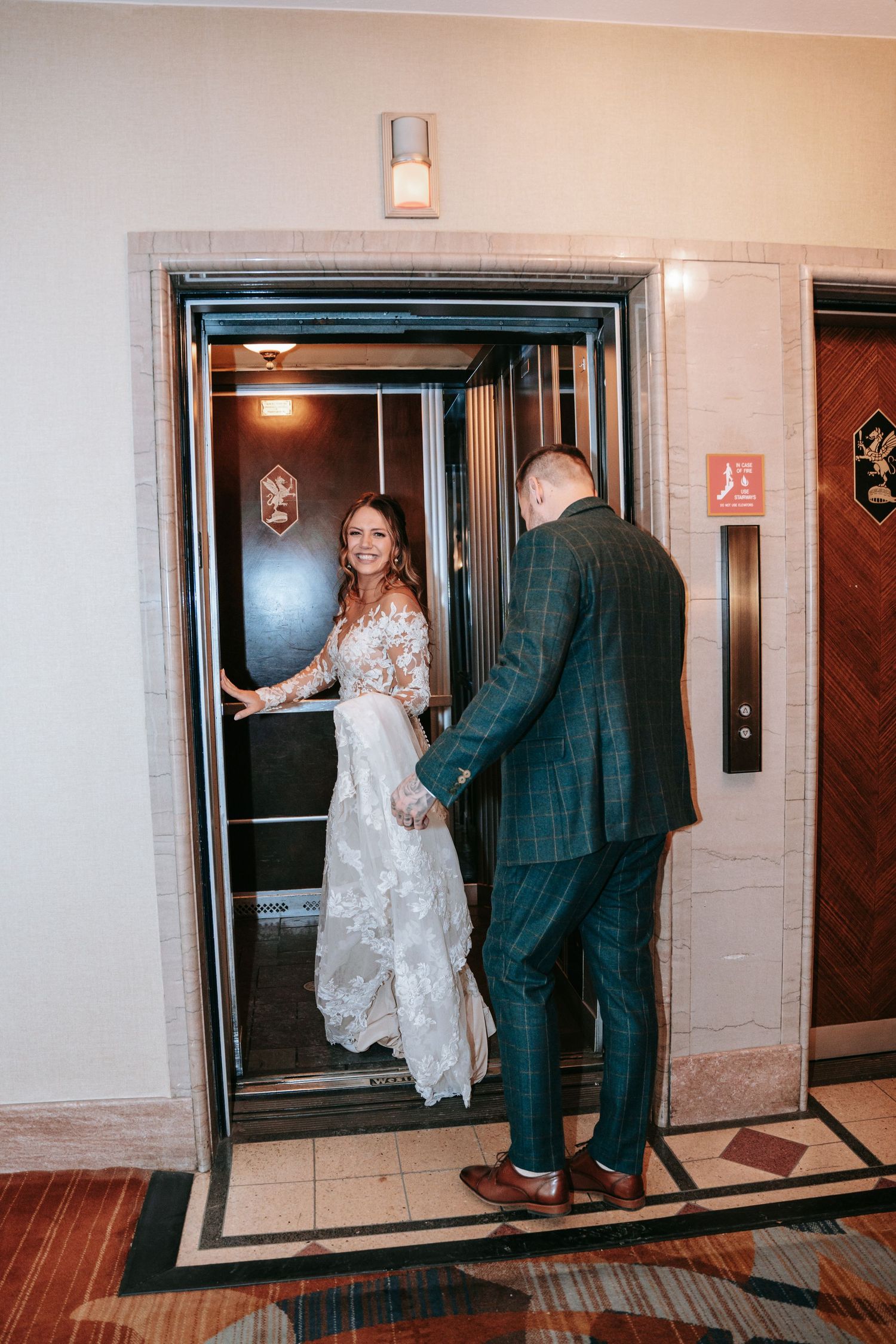 Smiling bride in lace gown holds elevator door open as groom in plaid suit steps inside.
