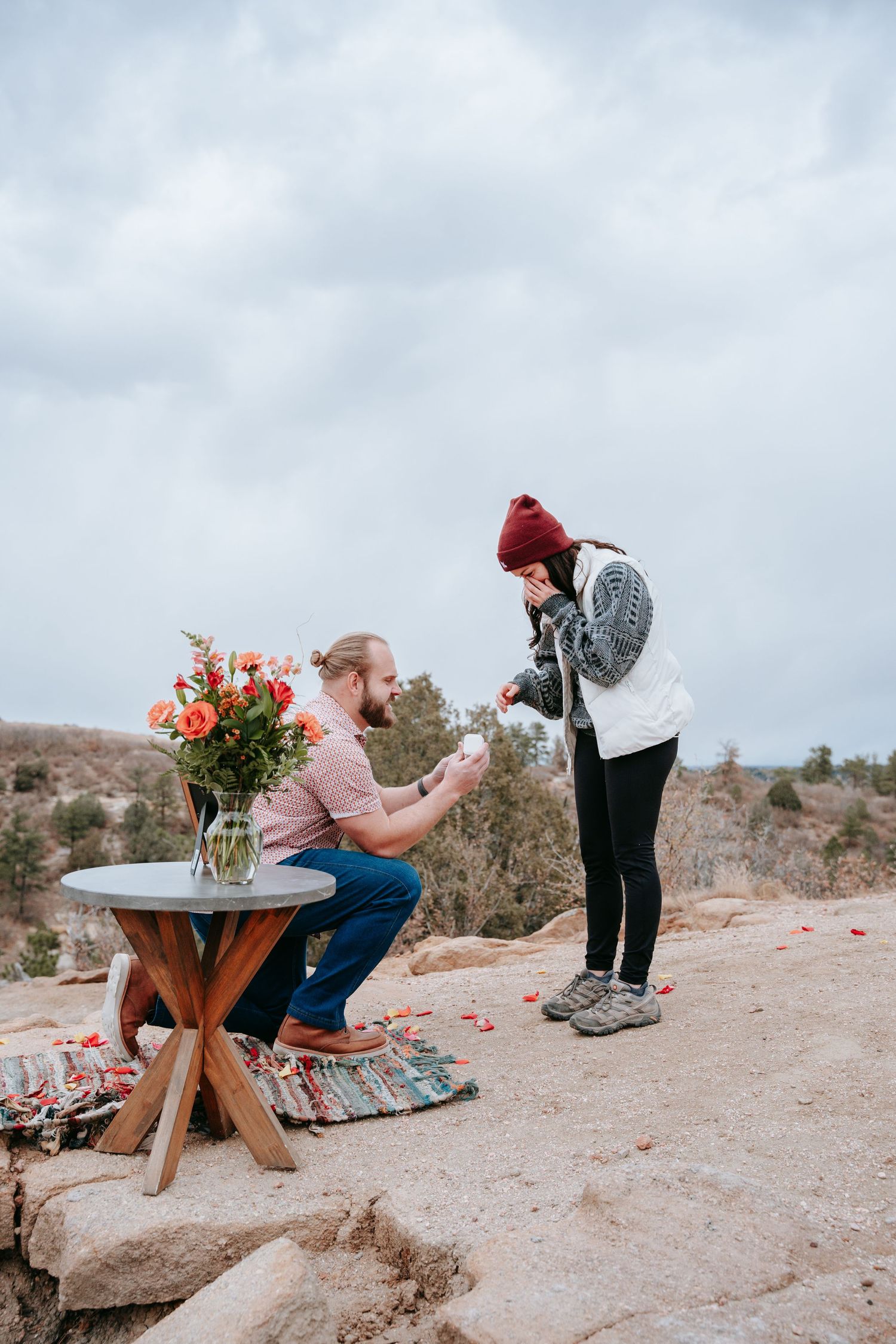 Man proposing to woman on rocky overlook with roses and rustic table in overcast outdoors.