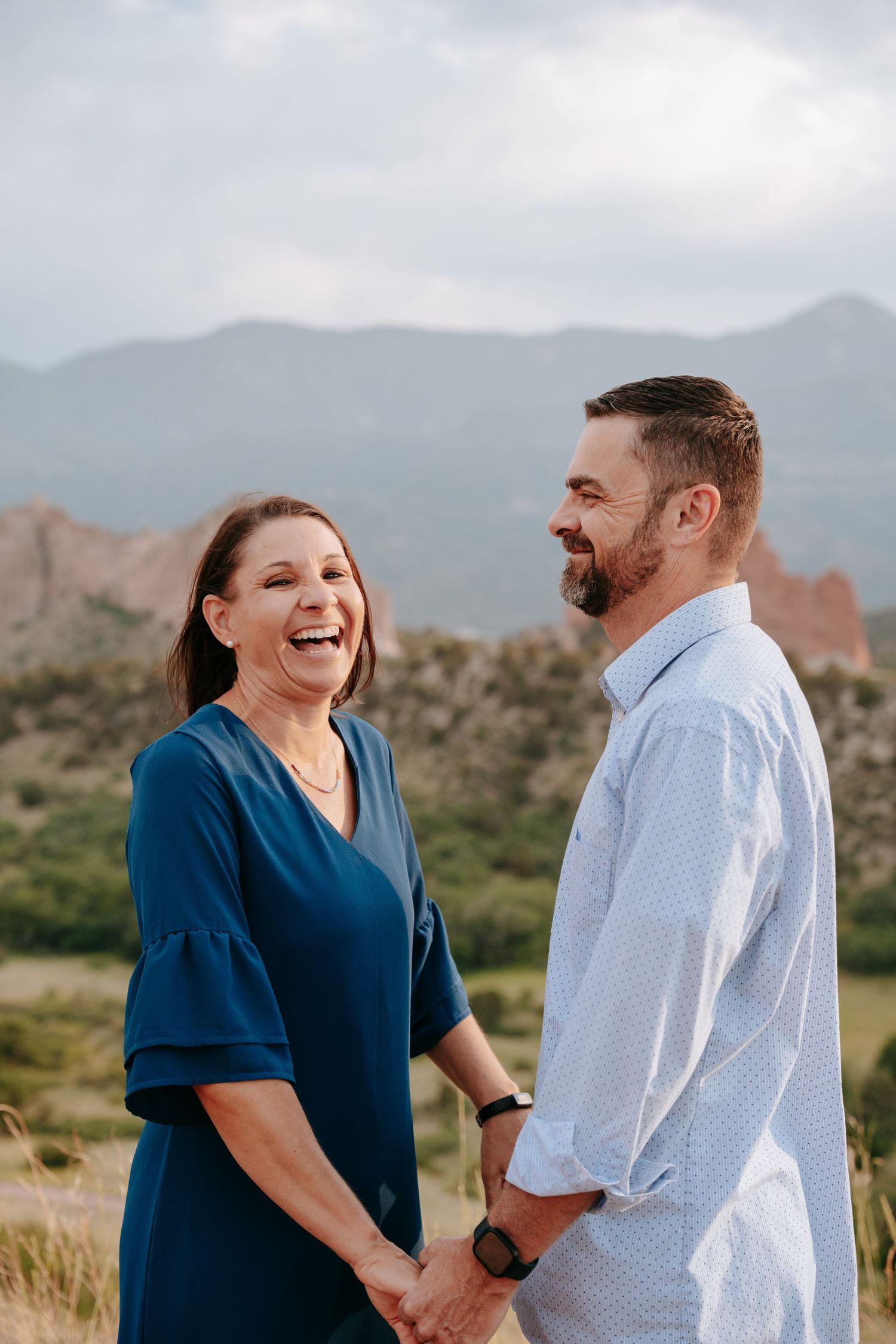 Laughing couple holding hands in a grassy field with Colorado mountains and red rocks behind.