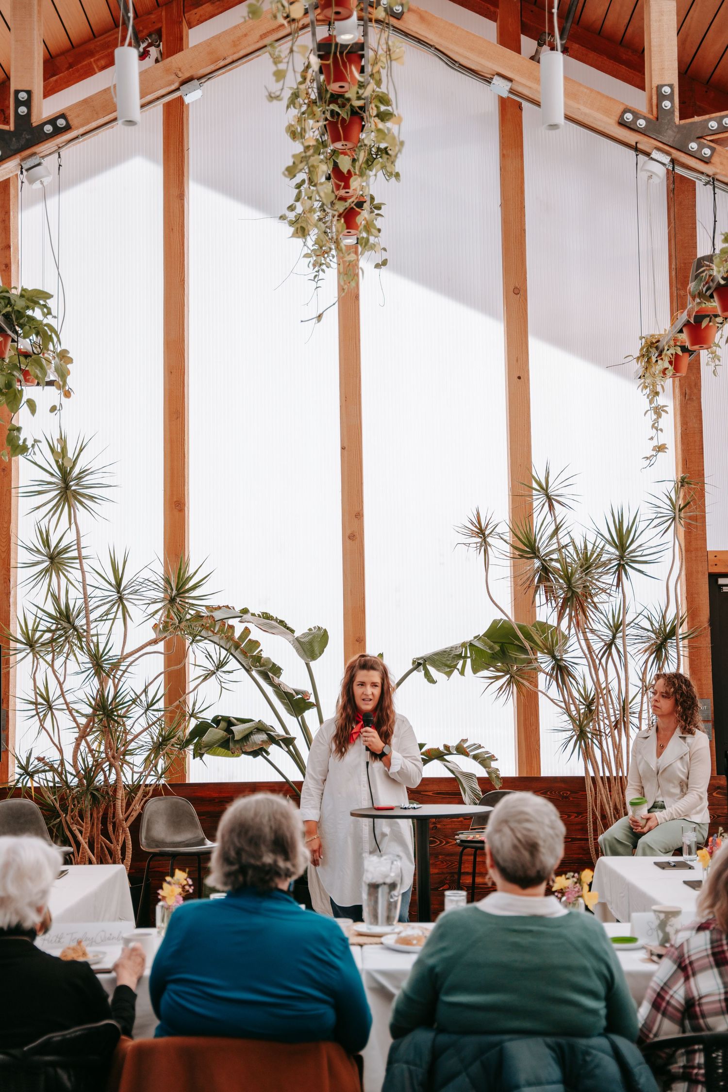 Woman speaking at elegant indoor event venue with large windows, tropical plants, and floral ceiling decorations.