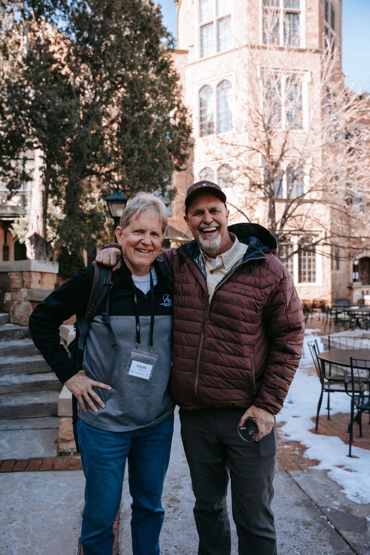 Two smiling adults posing outdoors in winter coats in front of a historic brick building.