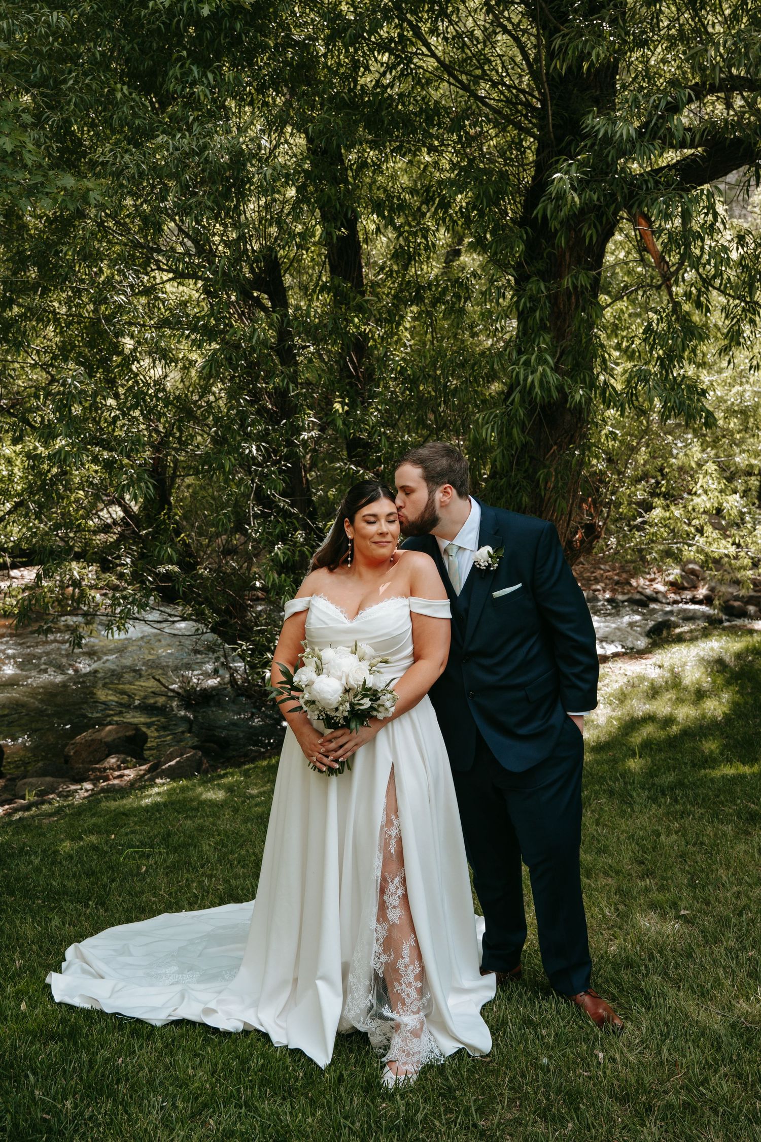Bride in off-shoulder gown holding white bouquet, groom in navy suit kissing her cheek, outdoors near creek.