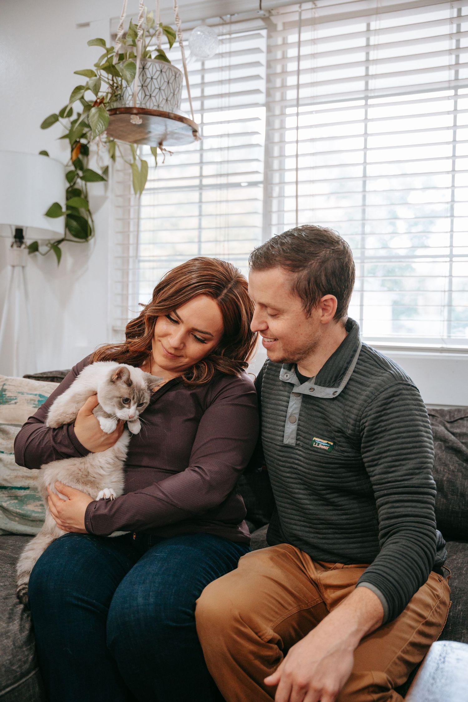 A smiling couple sits on a couch together, lovingly holding a fluffy Ragdoll cat at home.