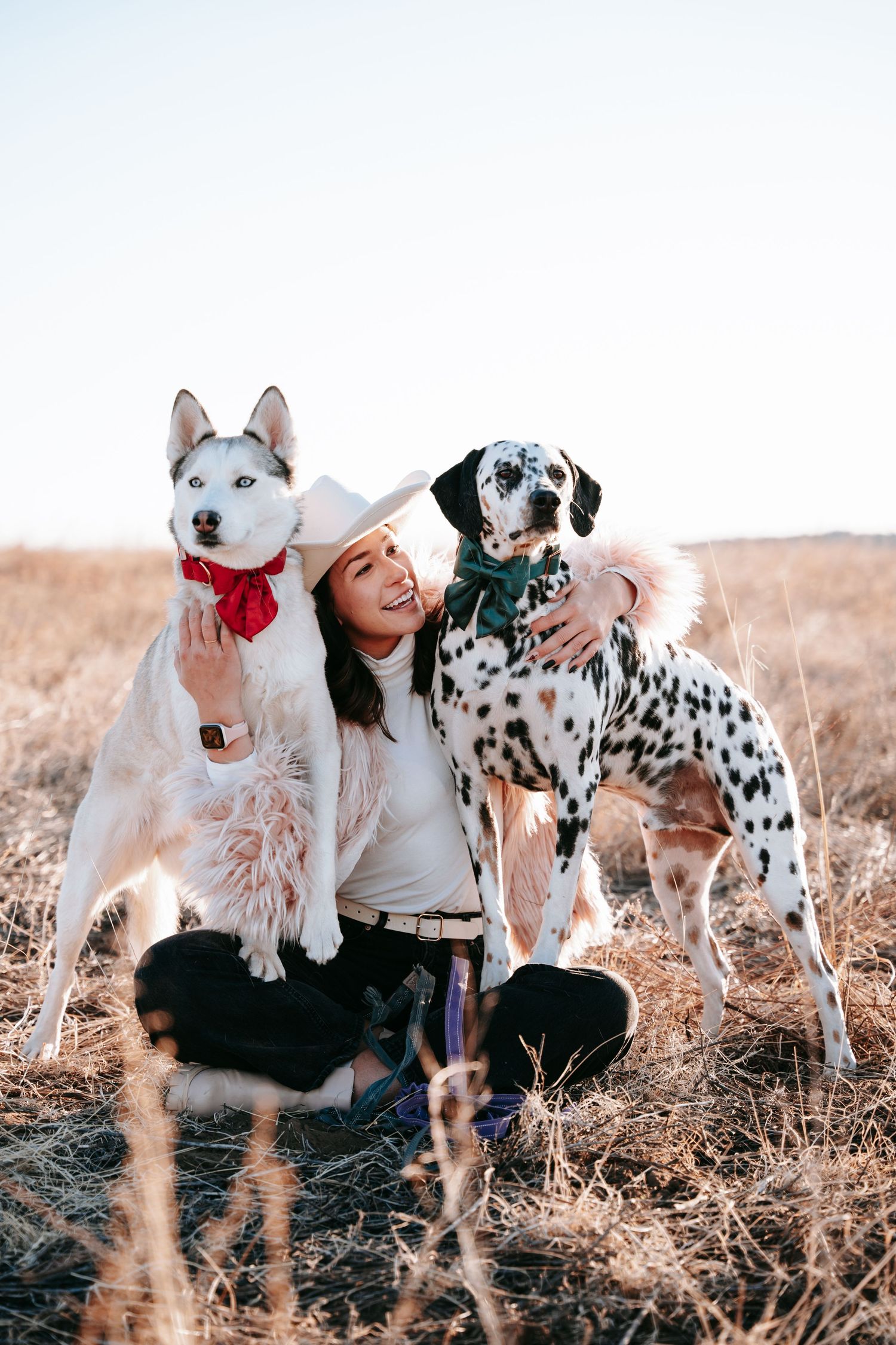 Happy woman in white cowboy hat posing with Husky and Dalmatian dogs in golden field.