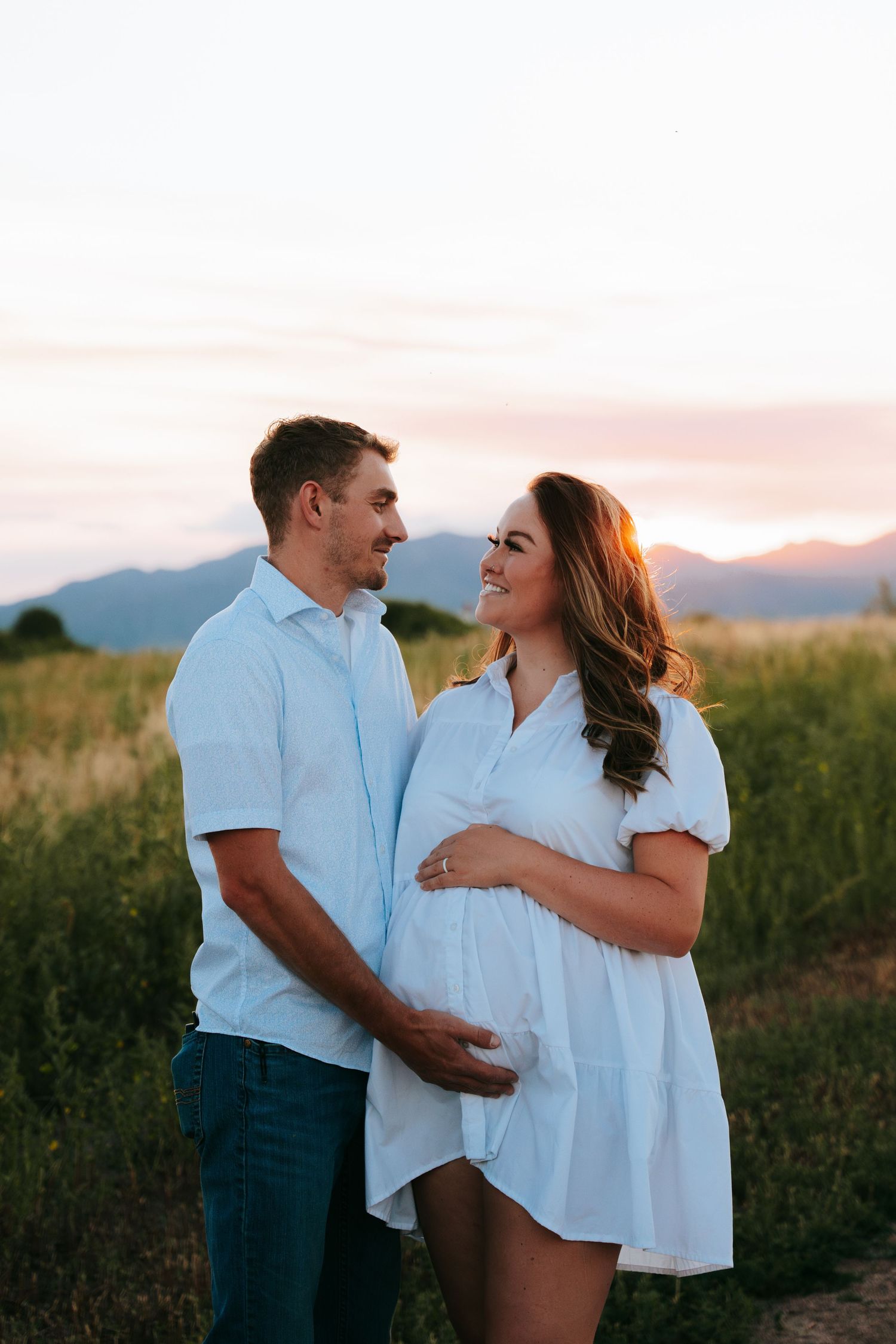 Expecting couple sharing a tender moment at golden sunset, woman in white dress cradling baby bump in mountain field.