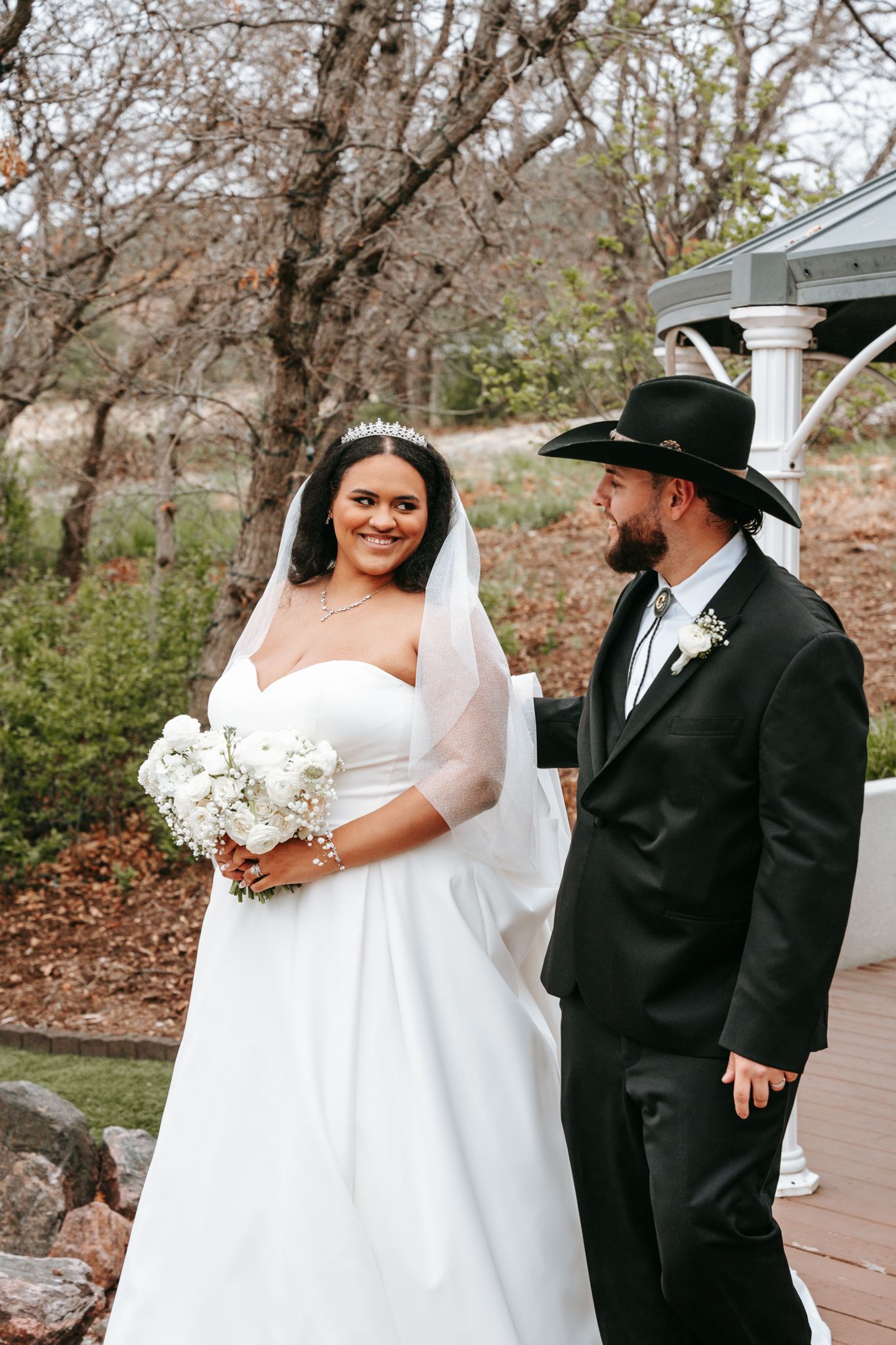 Bride in strapless gown with veil and groom in black suit with cowboy hat pose at garden gazebo.