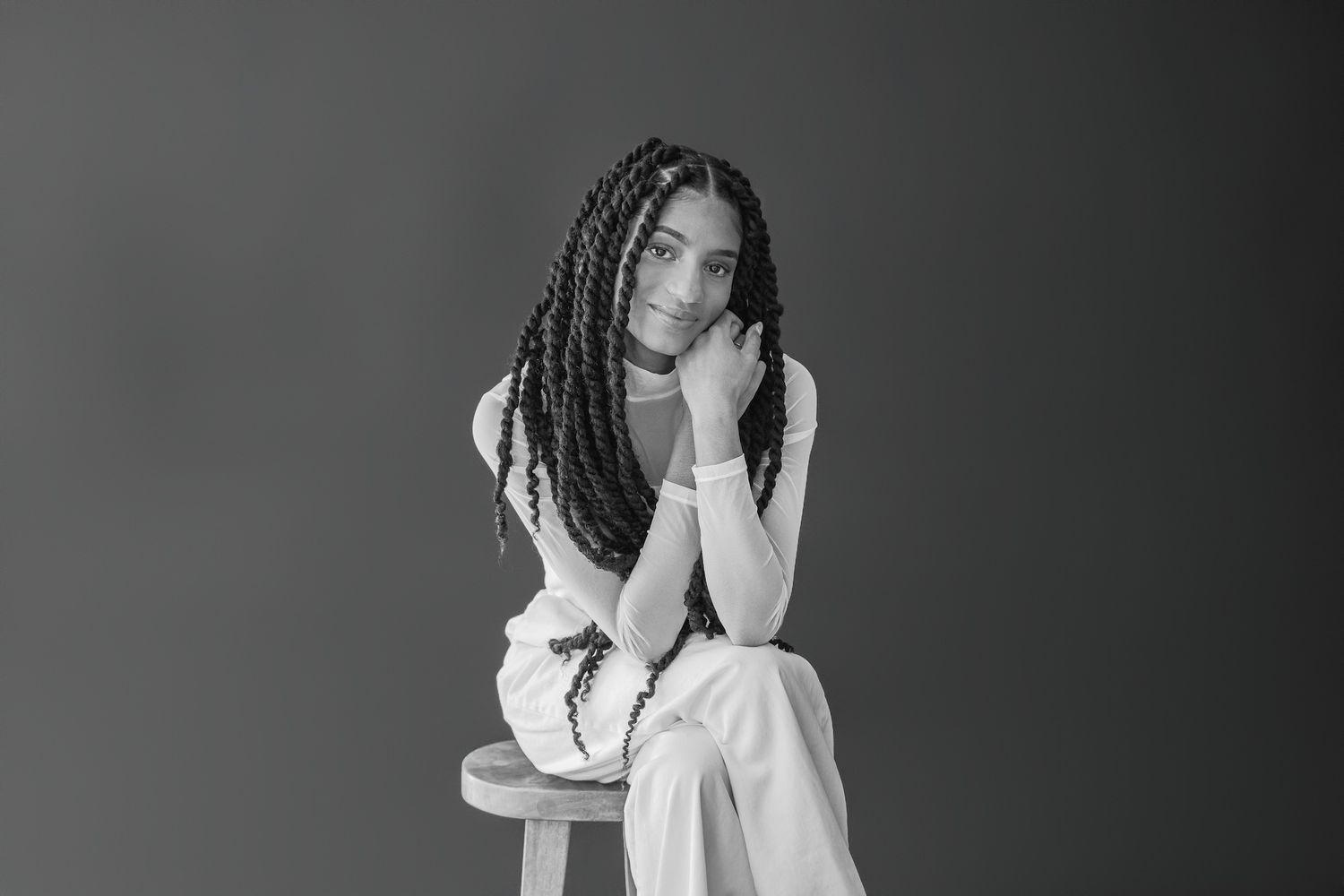 Young woman with long braids sitting on a stool in a black and white studio portrait.