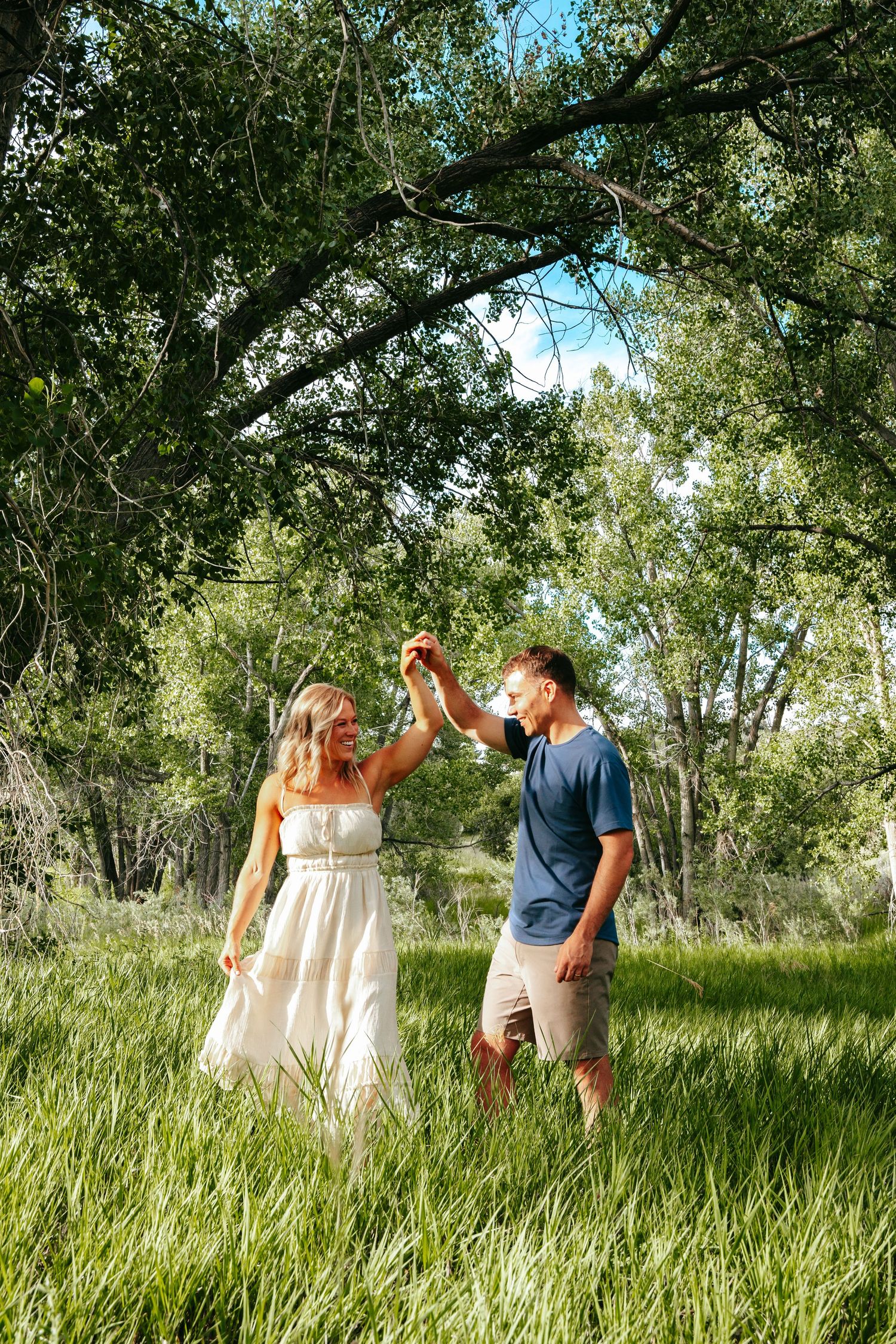Couple holding hands and dancing in lush green meadow surrounded by tall trees in sunlight.