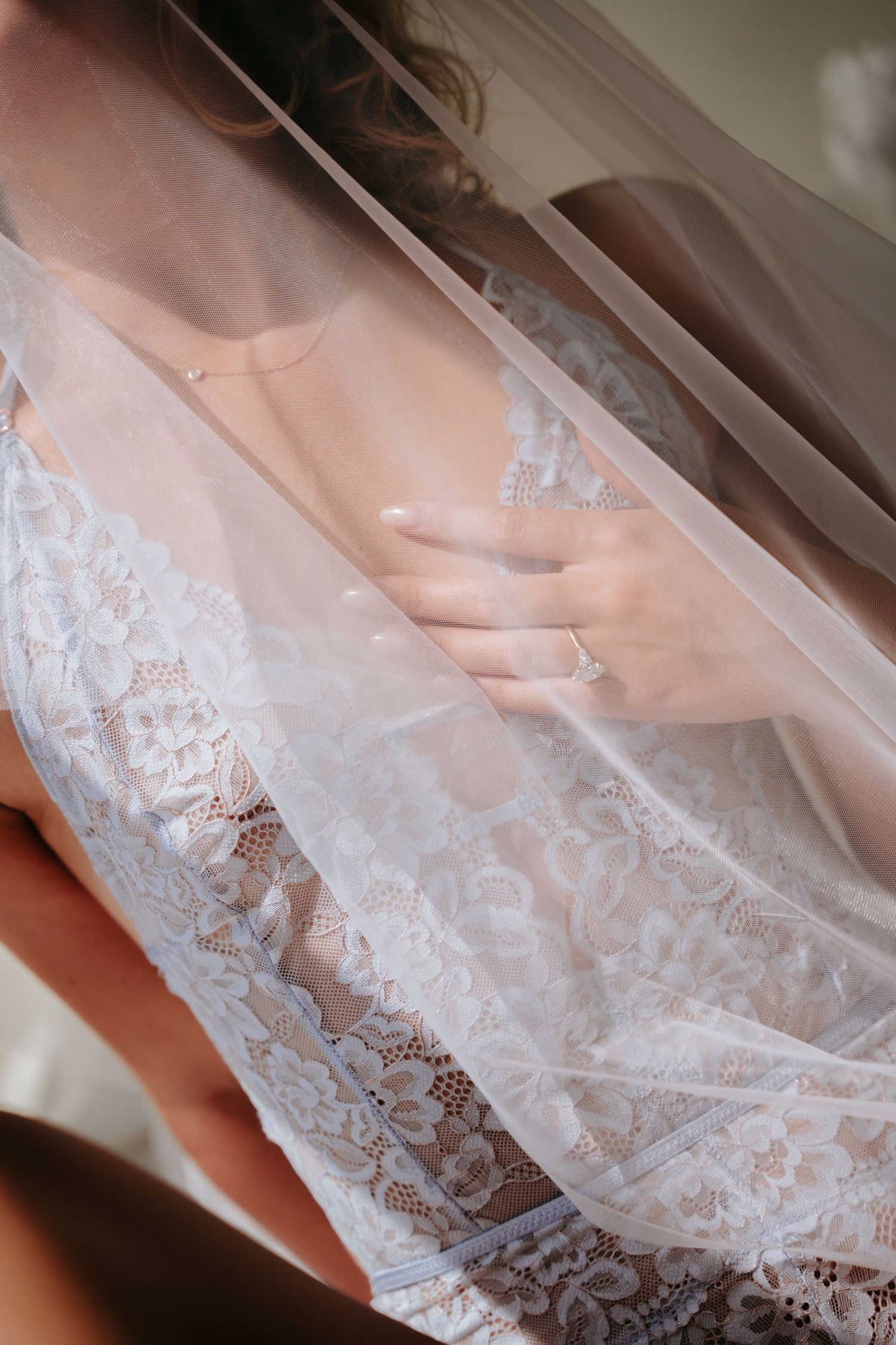 Bride in white lace wedding dress with veil draped over her, hand resting on chest showing elegant ring and necklace.