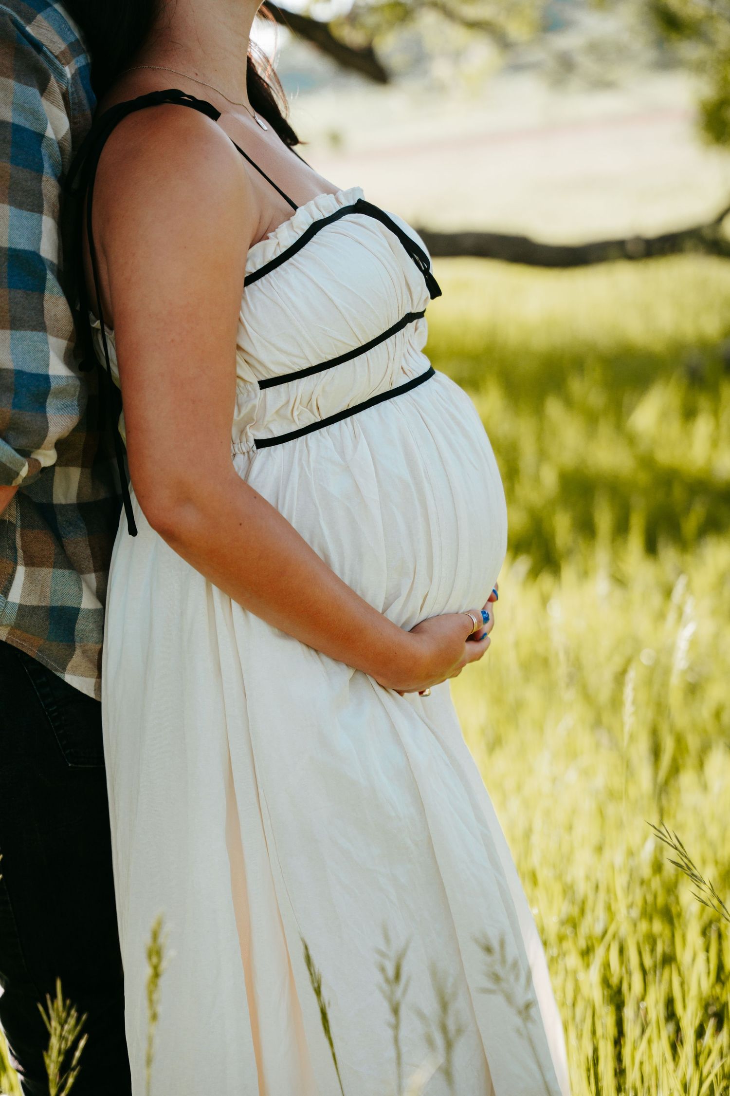 Close-up of pregnant woman in white dress gently holding baby bump while standing in sunlit green field.