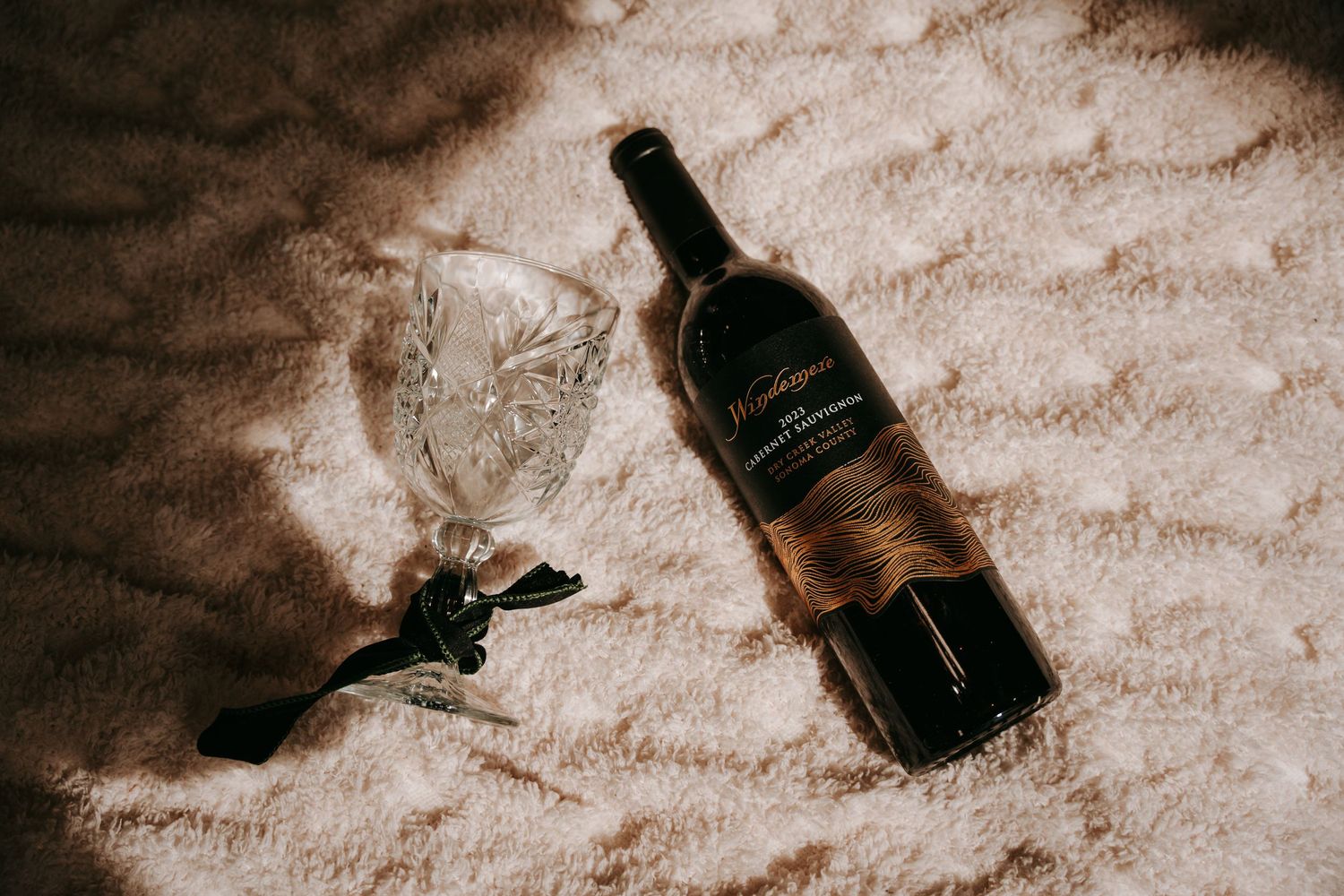Bottle of red wine with crystal glass lying on soft textured white carpet in warm sunlight.