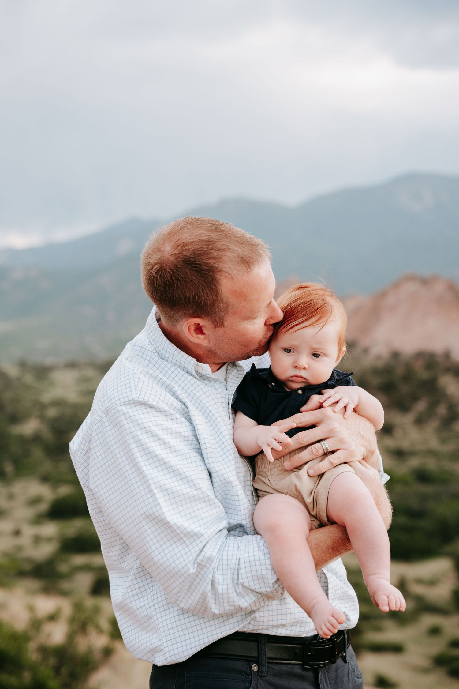 Father tenderly kissing red-haired baby boy while holding him outdoors with mountains behind.