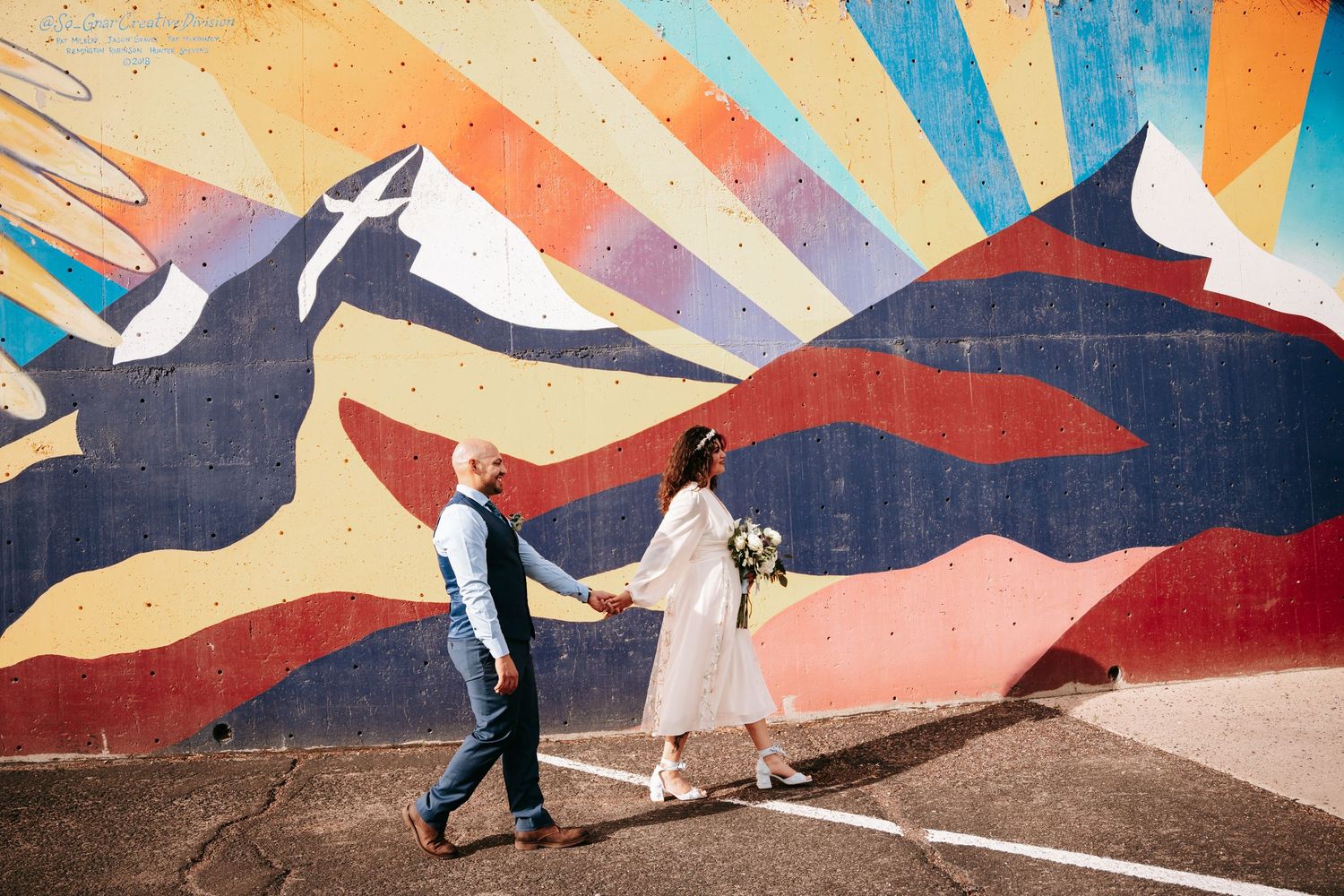 Newlyweds walking hand in hand past a vibrant colorful mountain mural on wedding day.