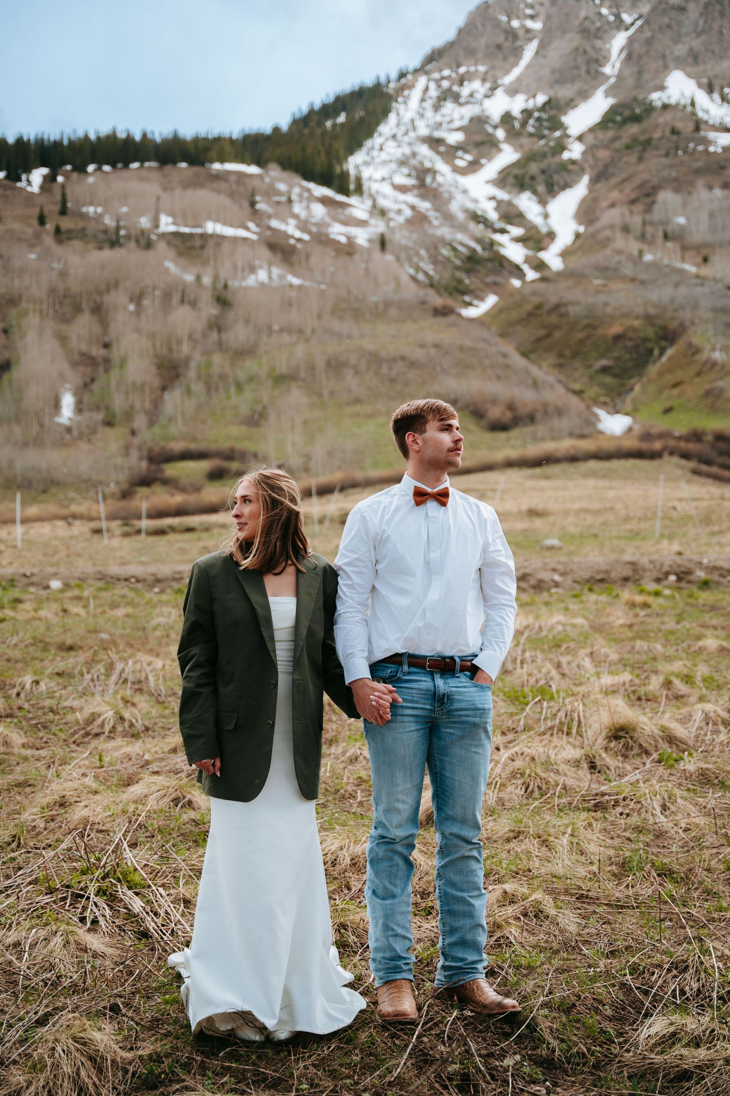 Wedding couple holding hands in mountain field, bride in white dress with green jacket, groom in jeans.