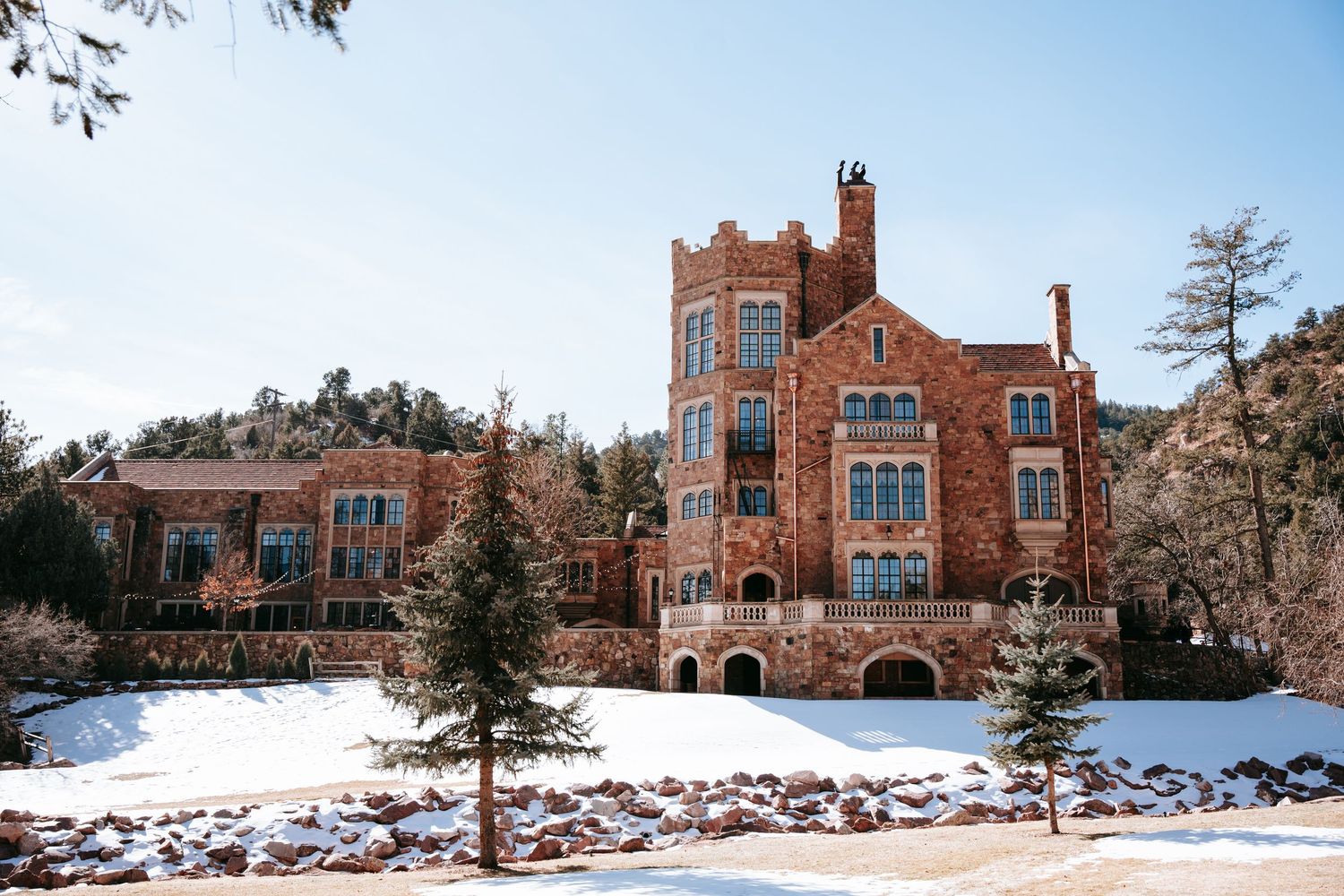 Historic red brick castle-style building surrounded by snow-covered grounds under a clear blue sky.