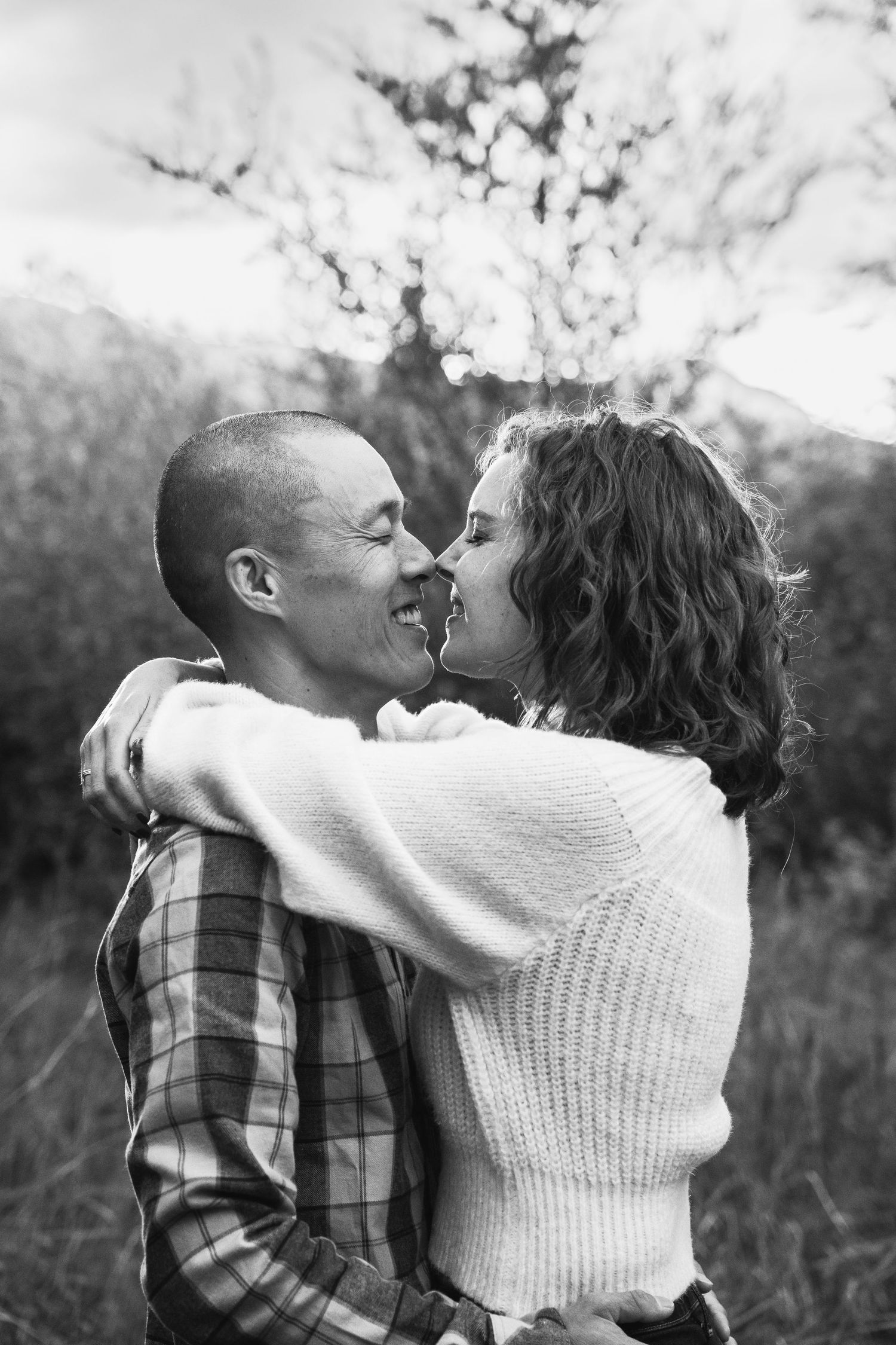 Black and white photo of a smiling couple embracing nose-to-nose in an outdoor field.