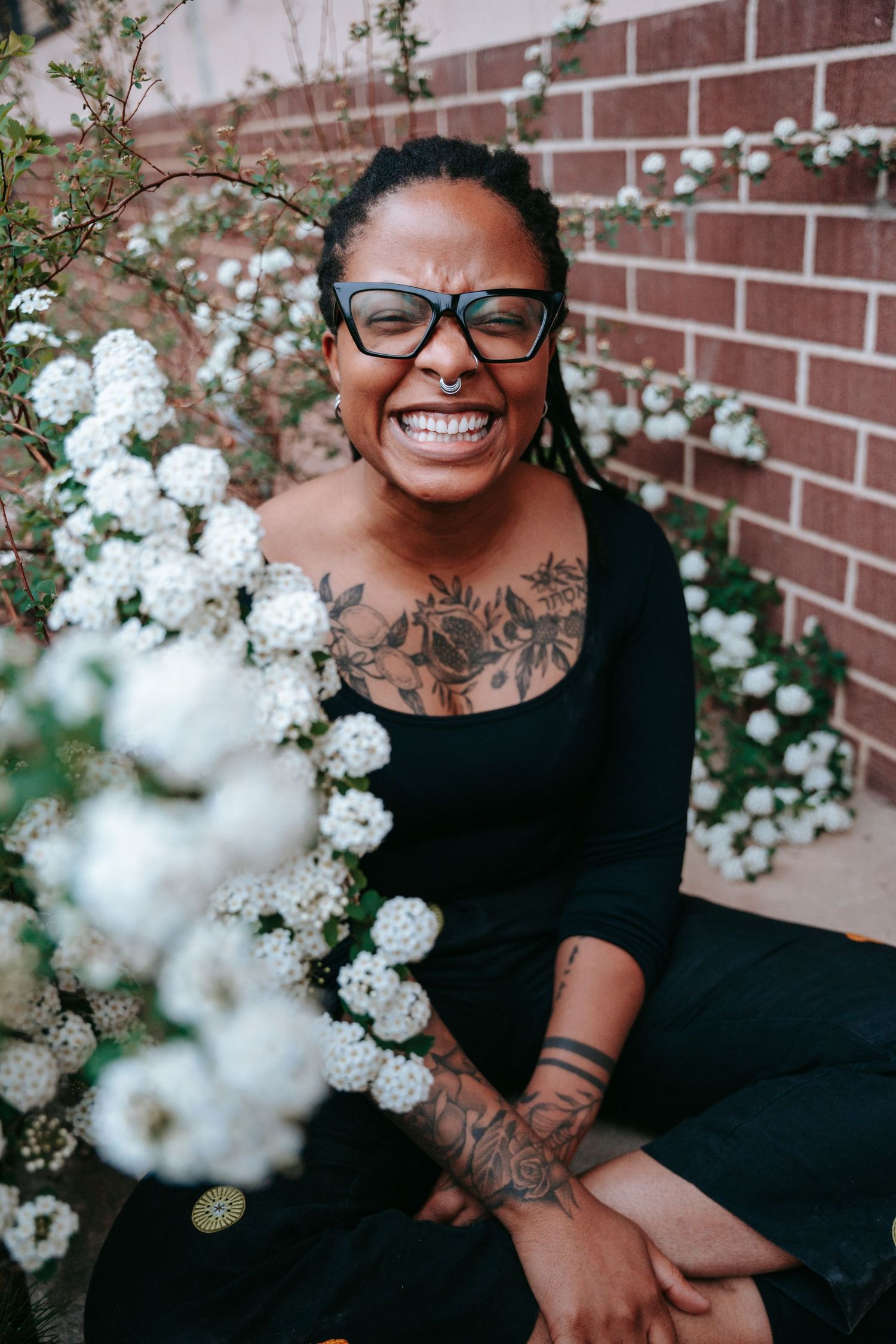 Smiling tattooed woman with dreadlocks and glasses sitting outdoors near white flowering bushes against a brick wall.