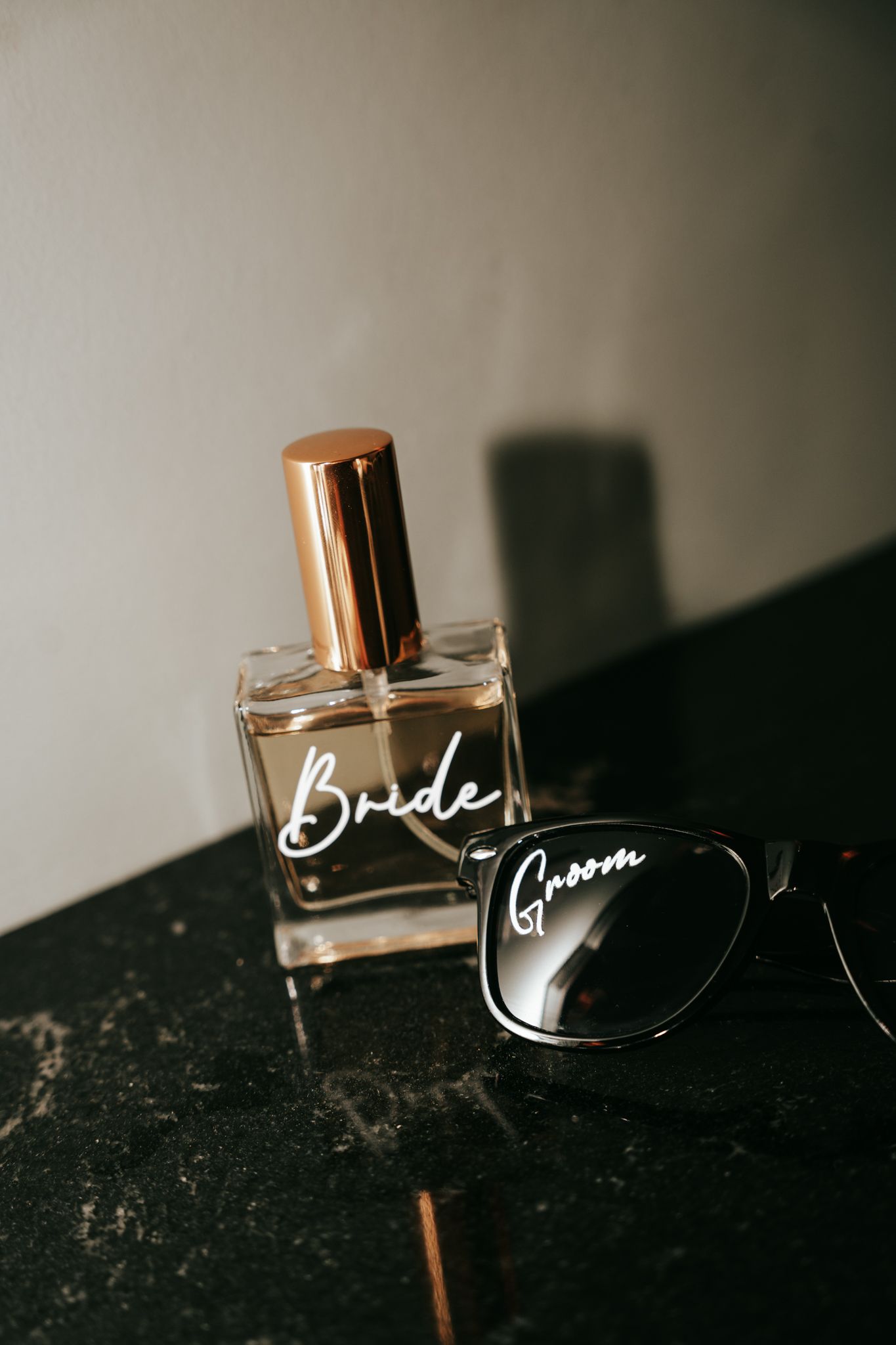 Wedding flat lay featuring a perfume bottle labeled Bride and sunglasses labeled Groom on dark surface.