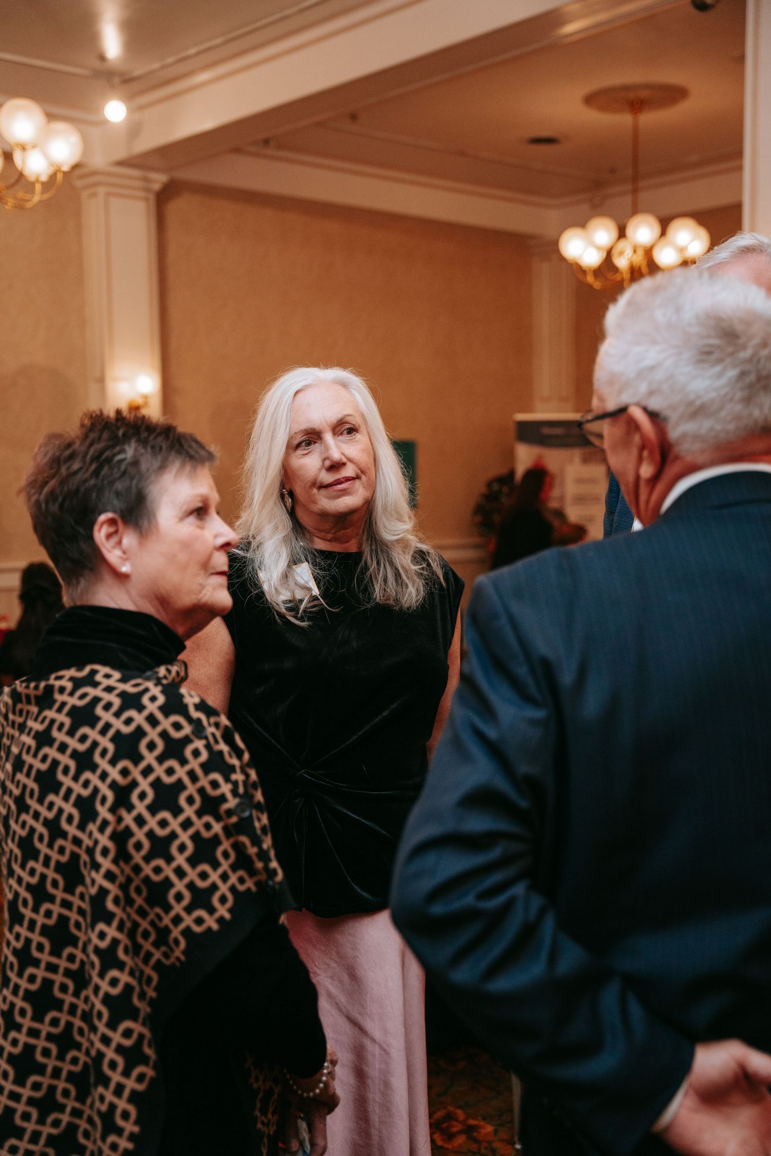Three guests chat at an elegant indoor reception with chandeliers, as a long-haired woman in black listens to a woman in a patterned jacket.