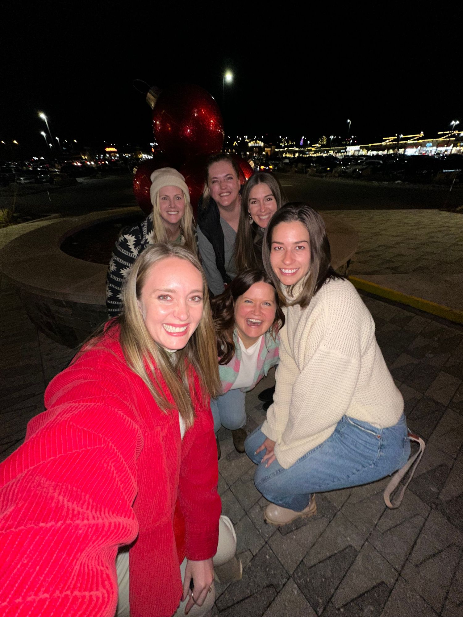 Group of six women smiling together for a nighttime selfie at an outdoor venue with city lights.