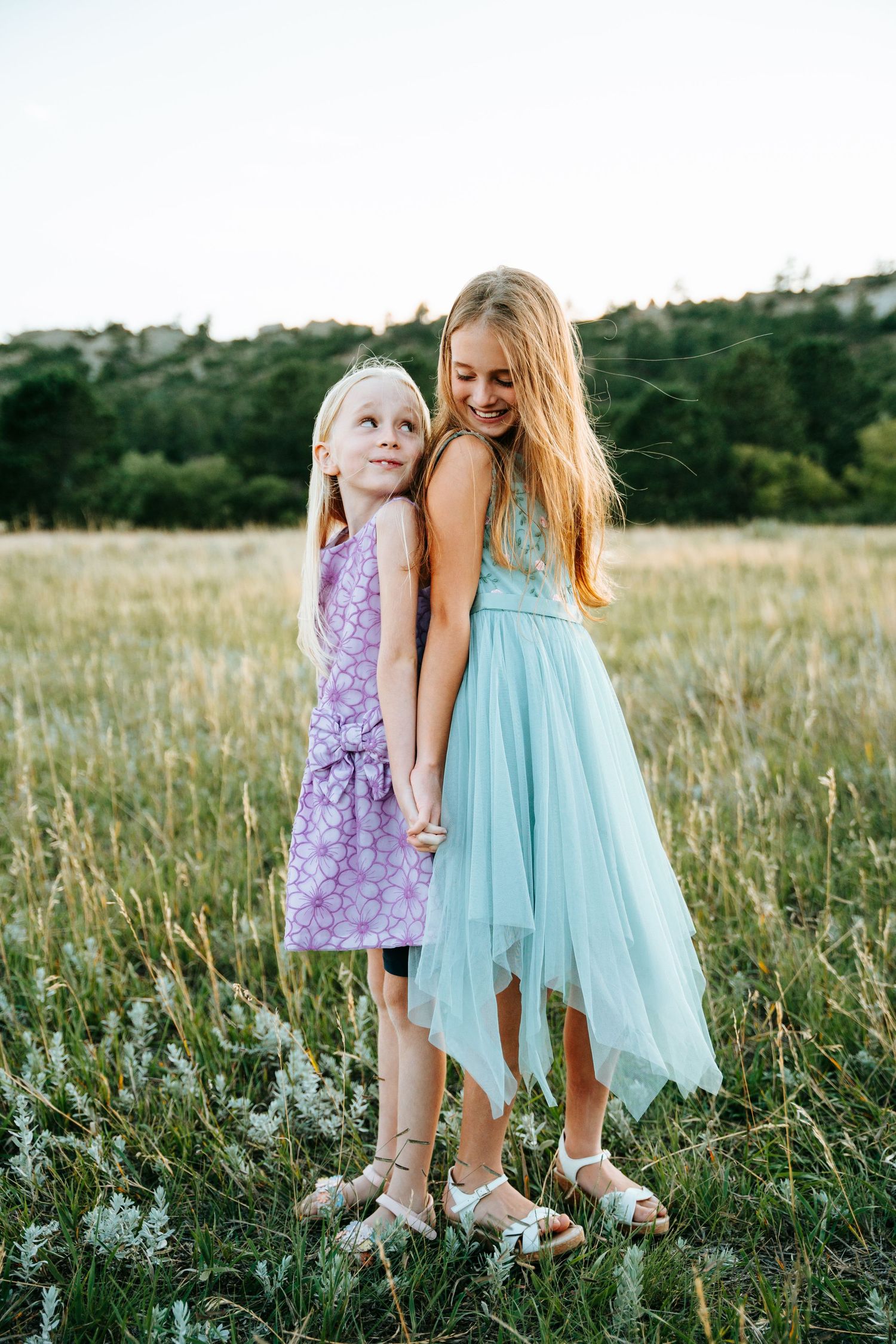 Two young blonde sisters in pastel dresses holding hands and smiling in a field.