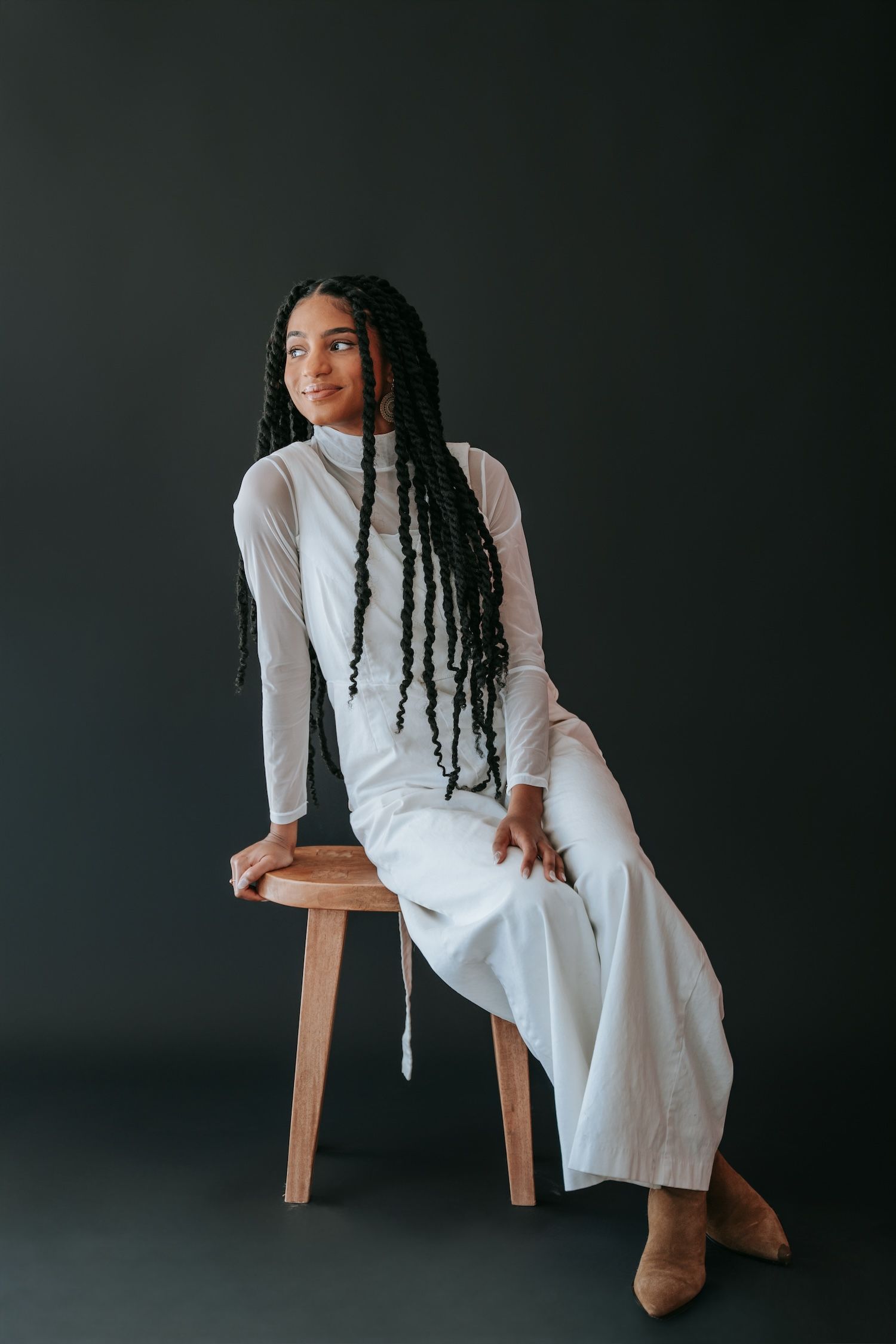 Elegant woman with long braids in white ensemble seated on wooden stool in studio.