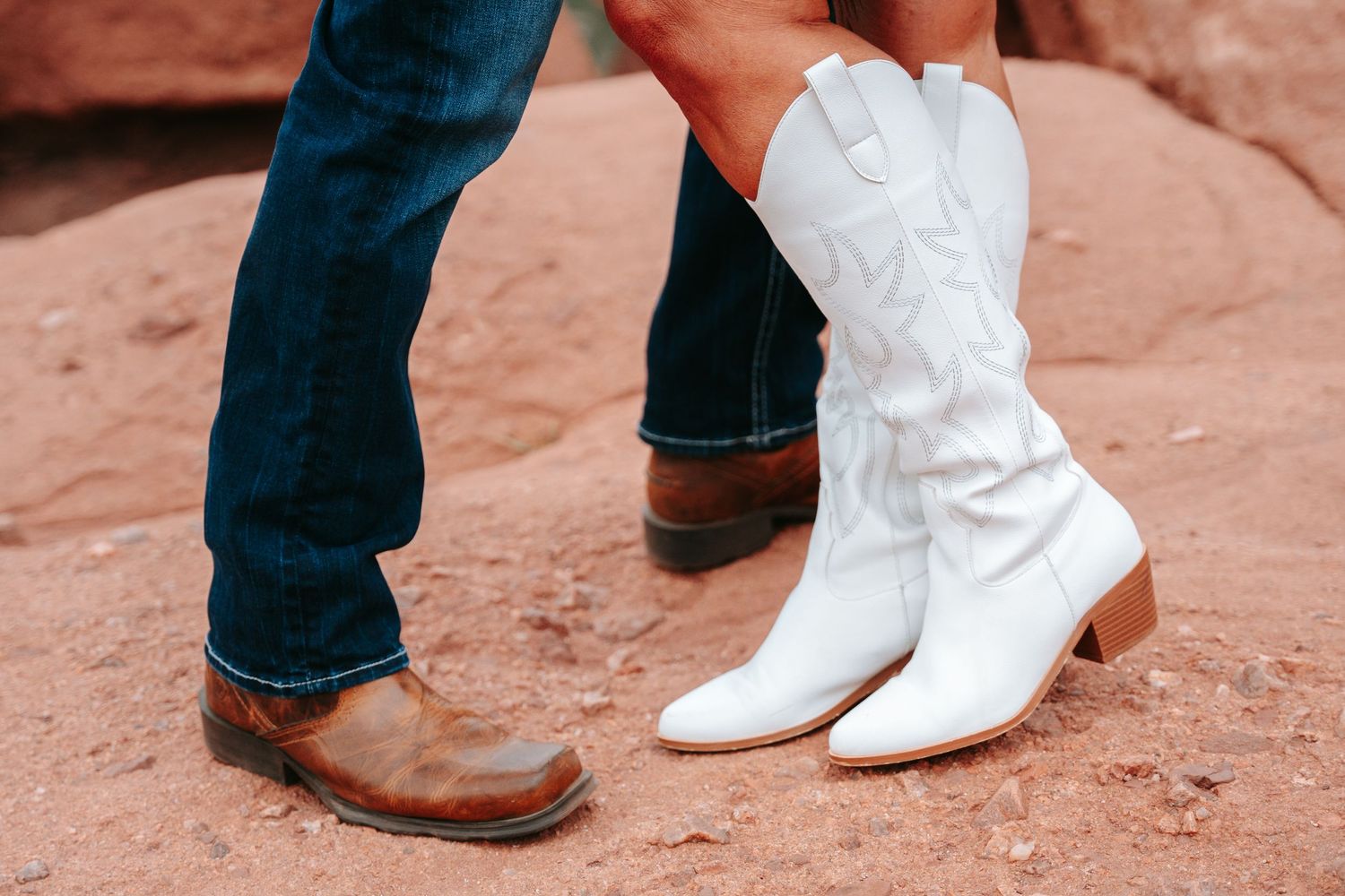 Close-up of couple wearing cowboy boots, brown leather and white western boots on red rock terrain.