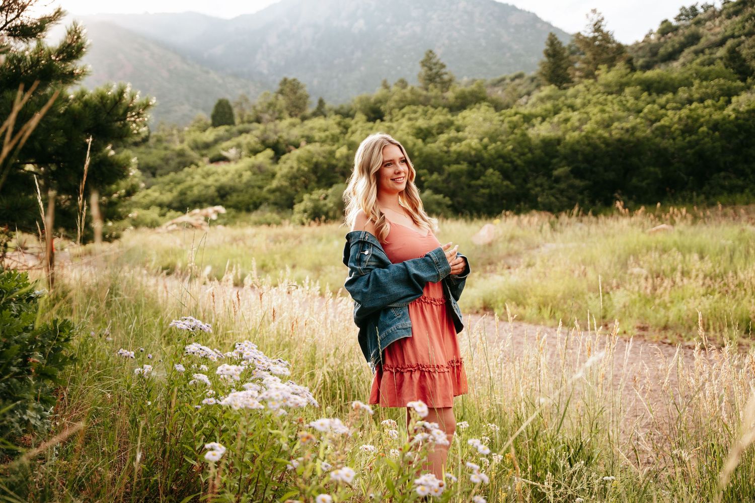 Blonde woman in coral dress and denim jacket standing in wildflower meadow with mountain backdrop.