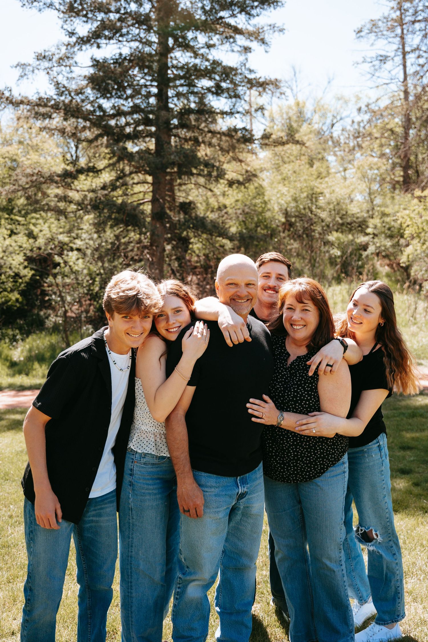 Smiling family of six hugging together outdoors in a sunny park with pine trees behind them.