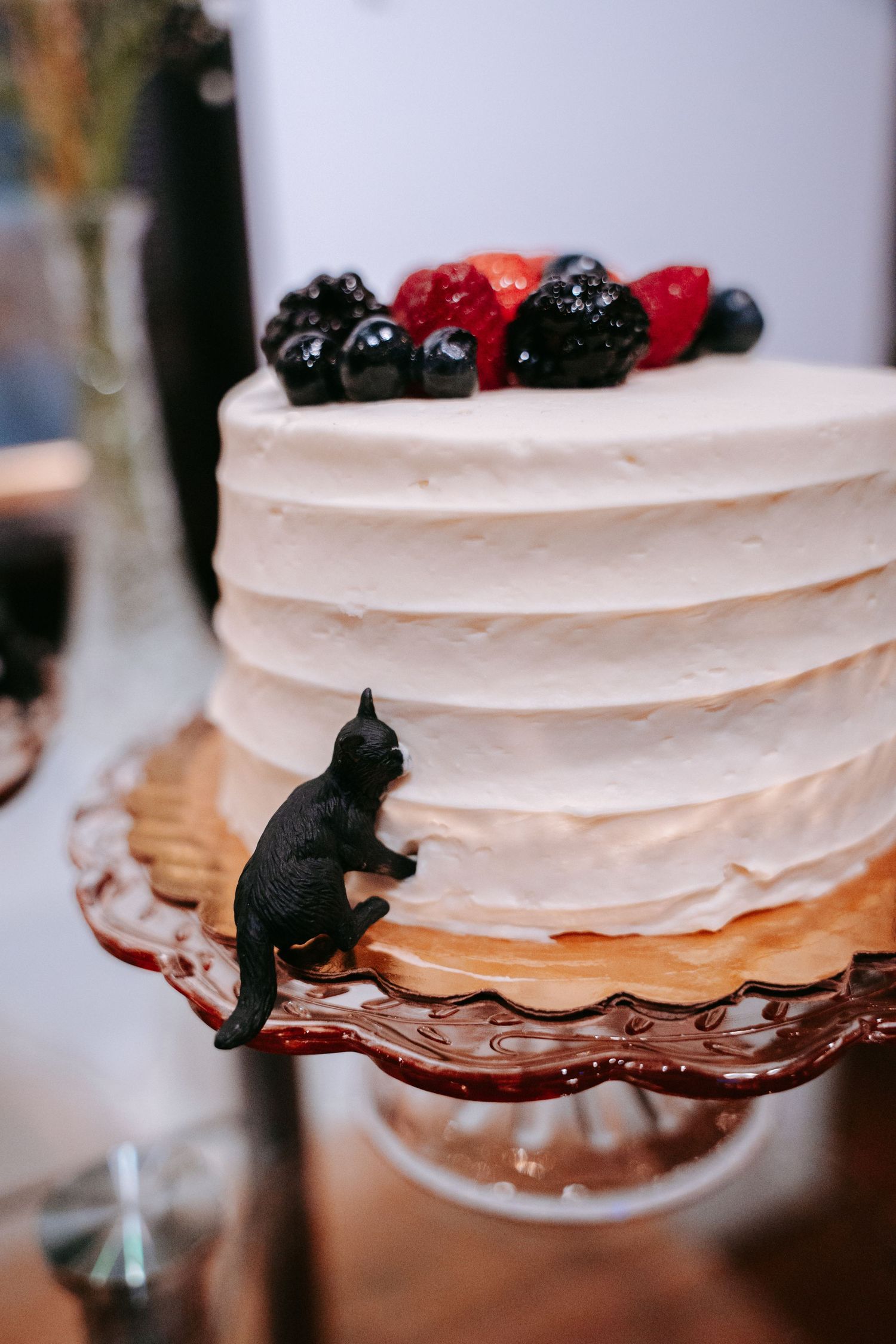 White layered wedding cake topped with fresh blackberries and a climbing black cat figurine.