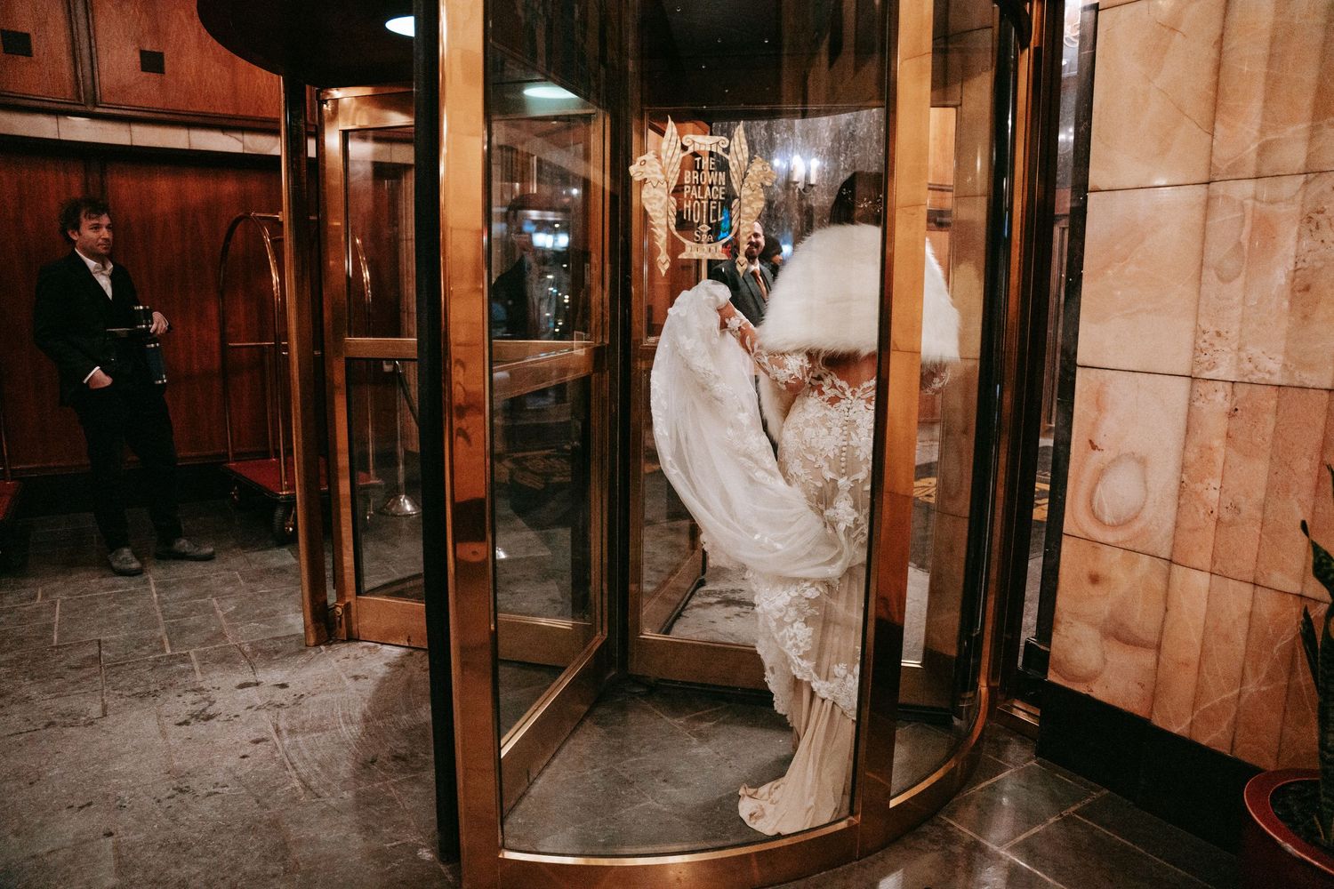 Bride in white lace gown enters a revolving brass door at an elegant wedding venue.