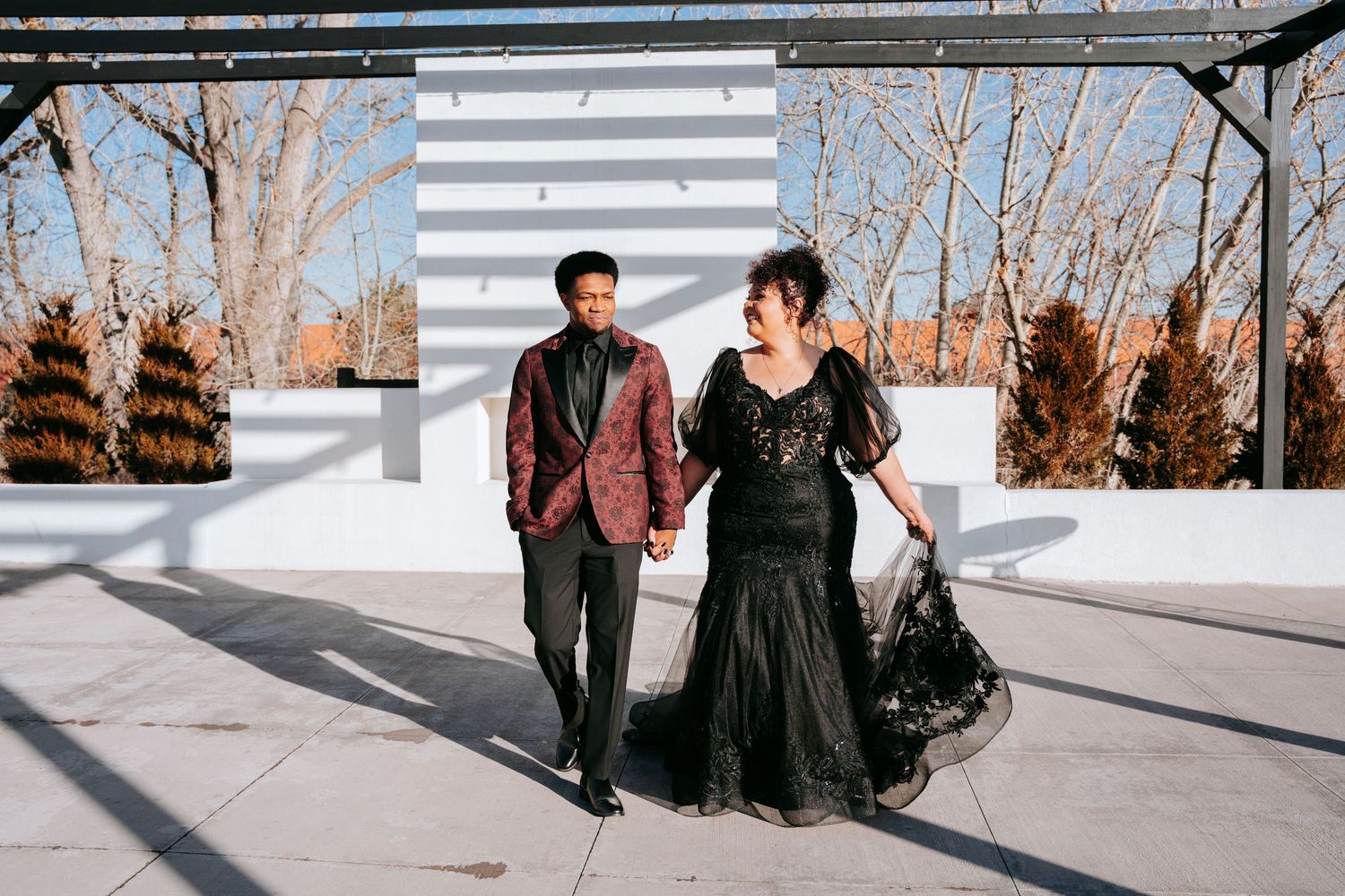 Couple in elegant black wedding attire hold hands walking on rooftop terrace in winter setting.