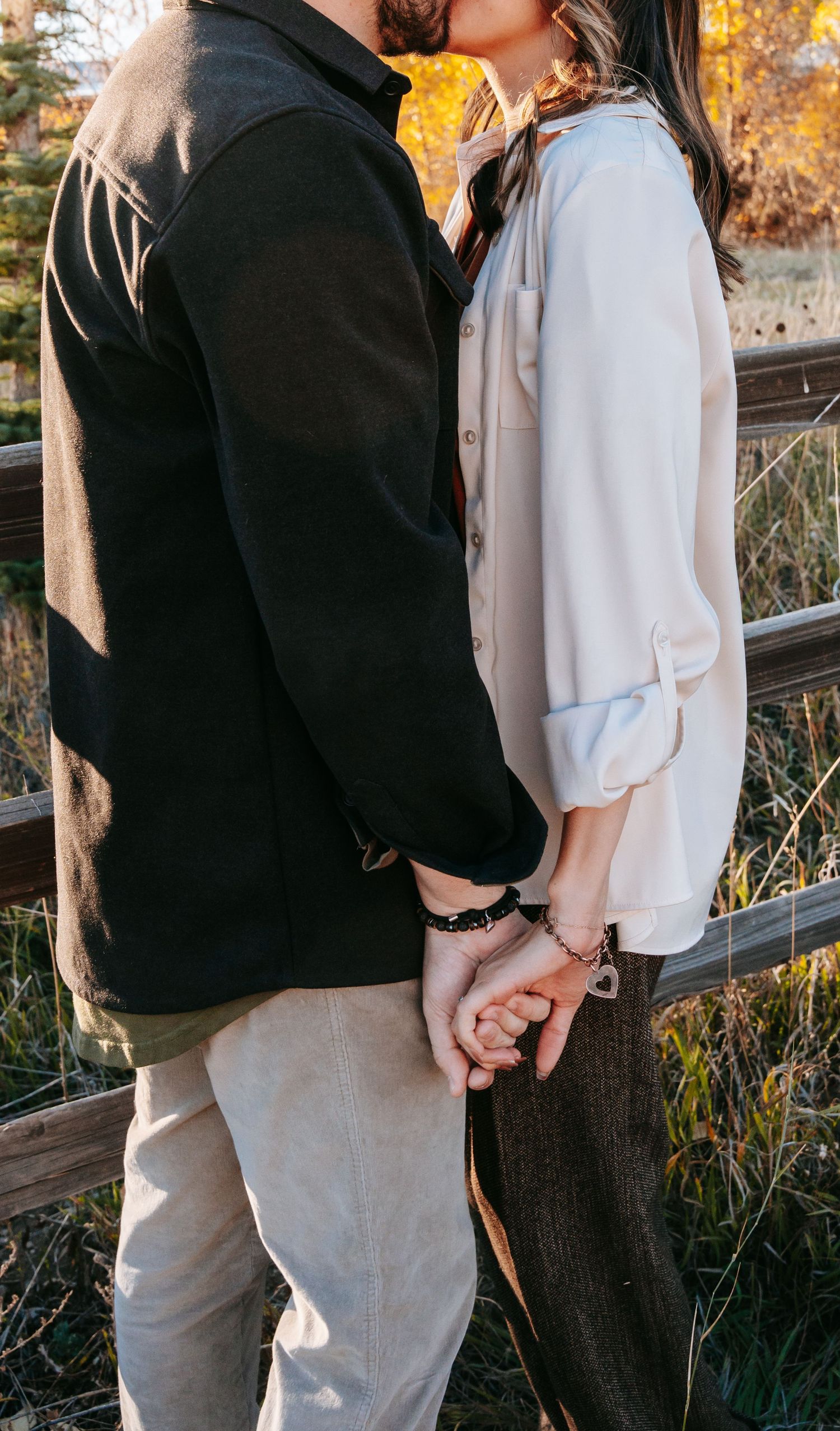 Close-up of couple holding hands and kissing by a wooden fence wearing matching bracelets.