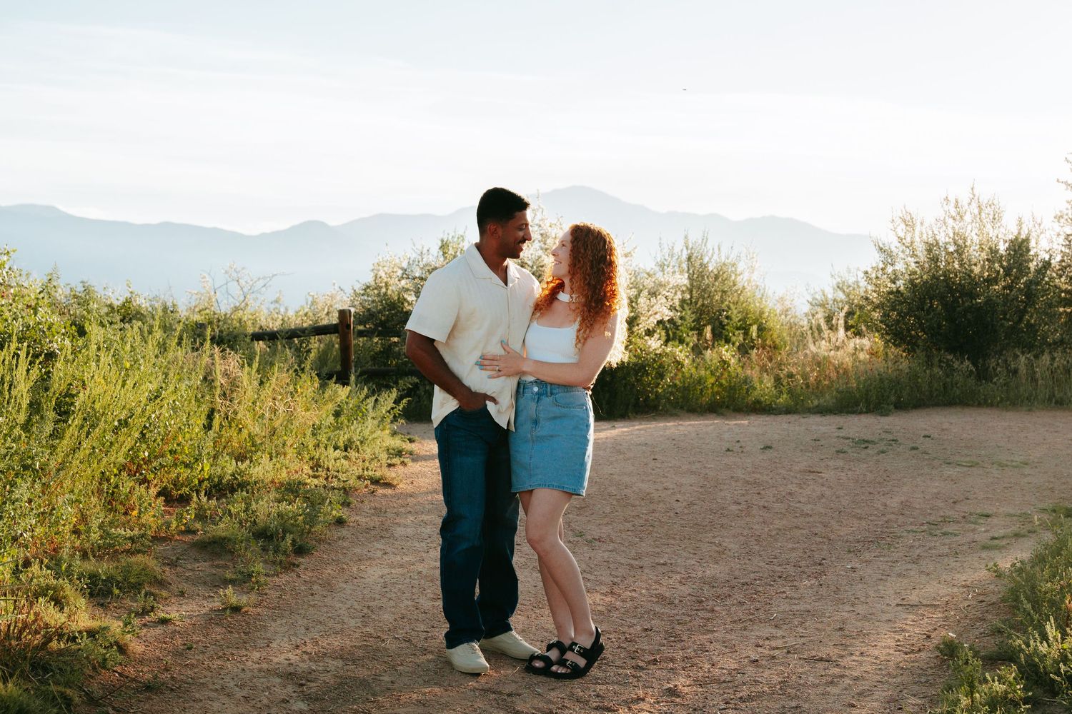 Couple standing together on a dirt path with mountains and greenery in the background during golden hour.