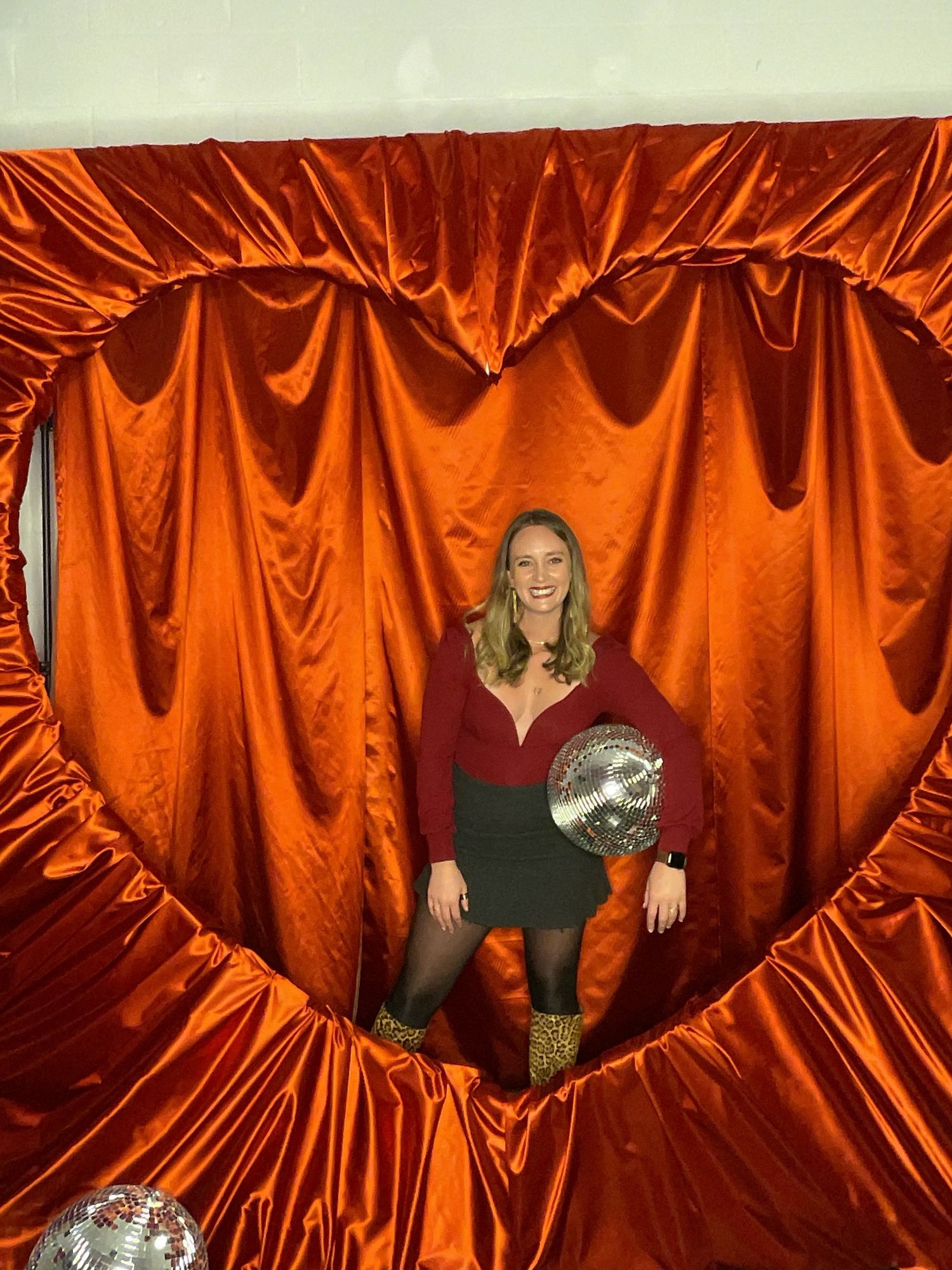 Woman smiling inside a red satin heart-shaped backdrop holding a disco ball at a Valentine's event.