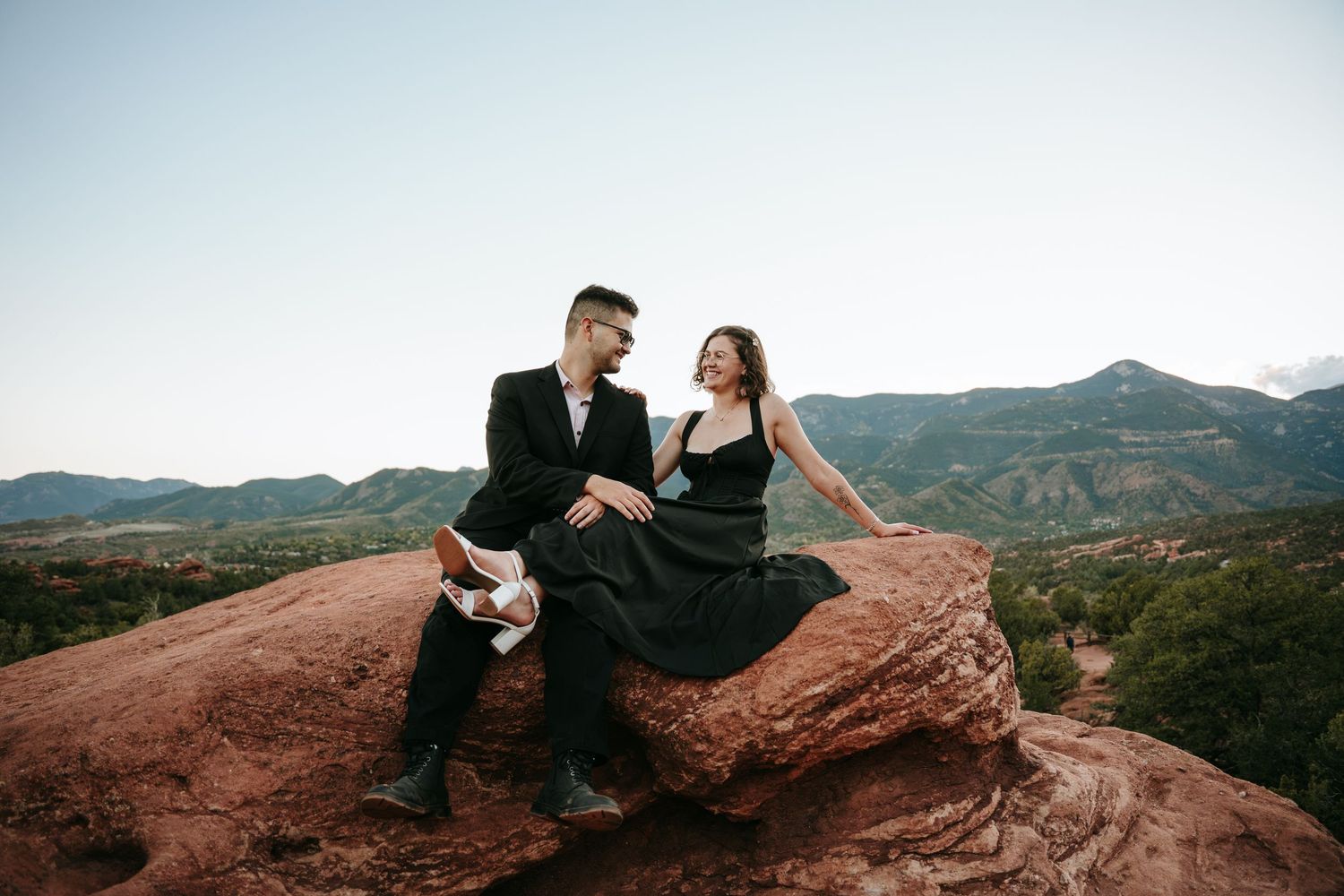 Couple in formal attire sitting close together on a red sandstone rock formation with green mountains and a clear sky behind them.