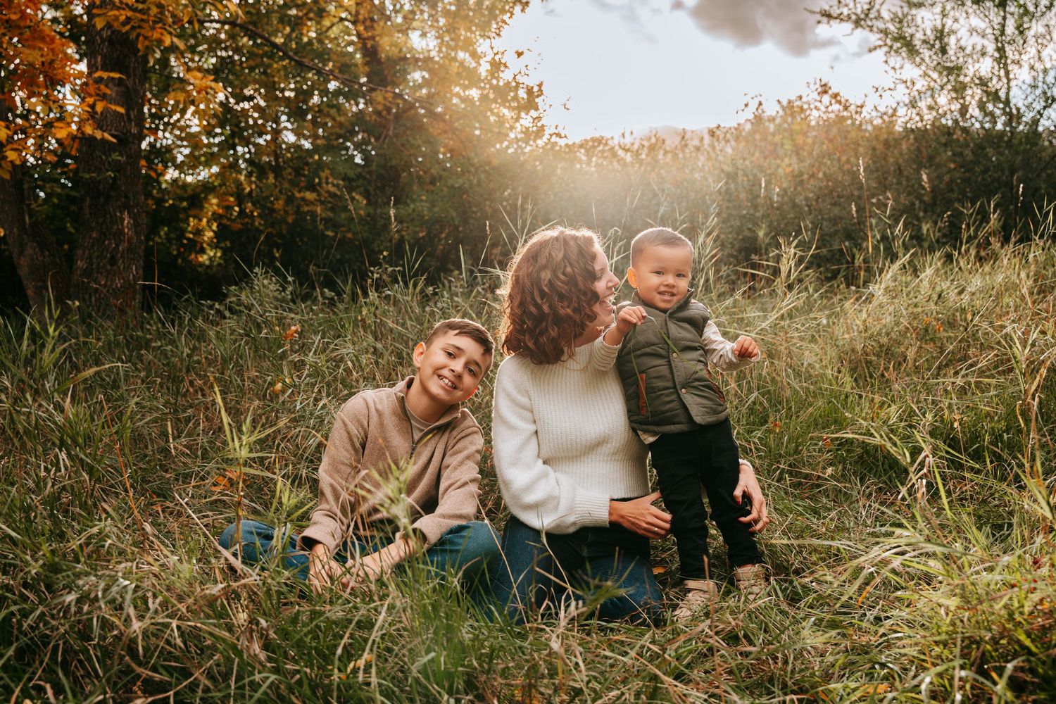 A mother sits in a sunlit autumn meadow with her two young children, surrounded by golden trees and tall green grass.