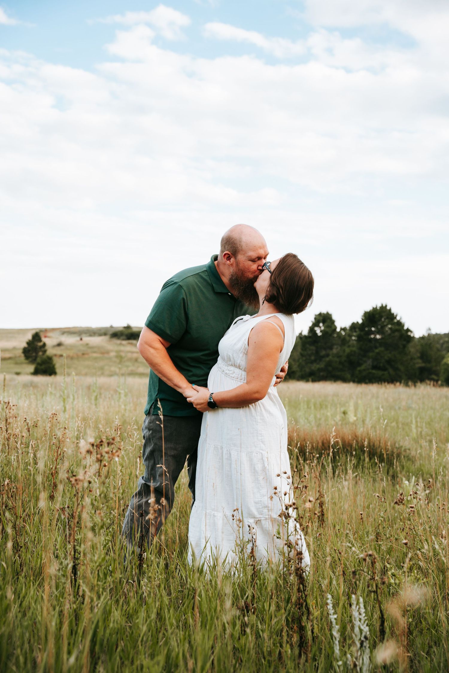 Romantic couple sharing a kiss in a wide open grassy field under a partly cloudy sky.