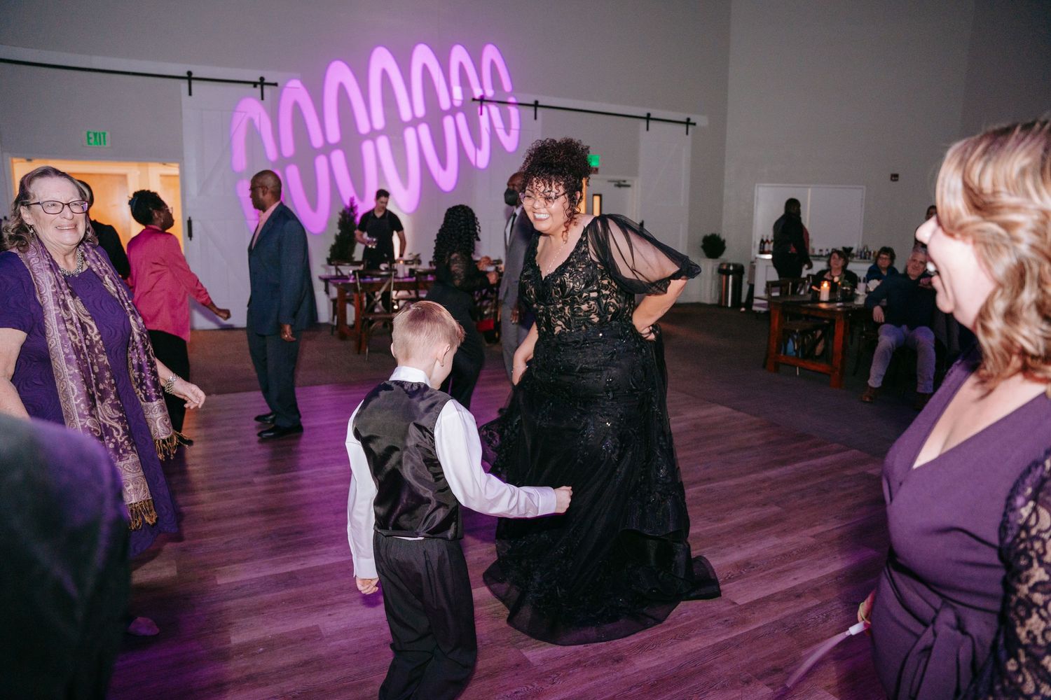 Woman in black lace gown dancing with young boy at wedding reception with purple neon lights.