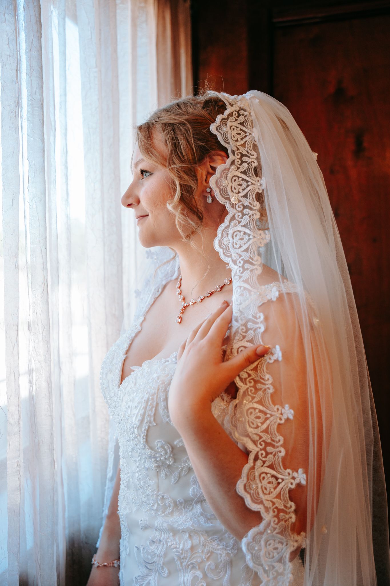 Smiling bride in lace wedding gown and ornate veil standing by window in soft natural light.
