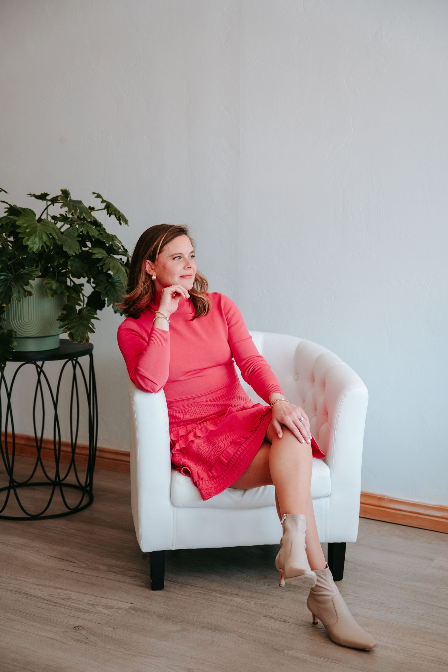 Woman in pink dress sitting thoughtfully in white armchair next to indoor plant.