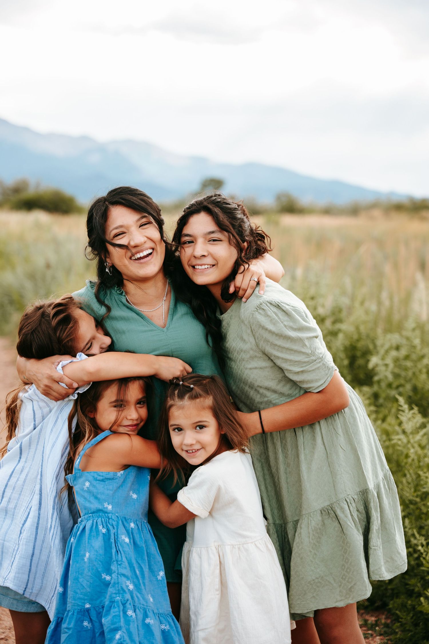 Joyful Hispanic mother laughing as four daughters embrace her in a green mountain meadow on cloudy day.