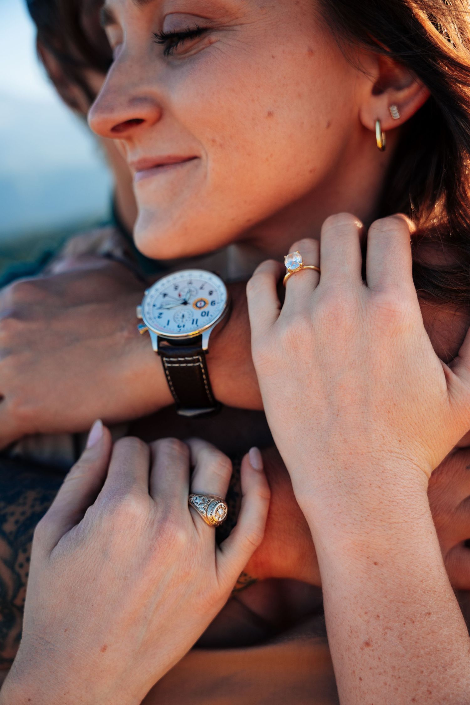 Close-up of smiling woman being embraced, showcasing oval diamond engagement ring and partner's vintage watch.