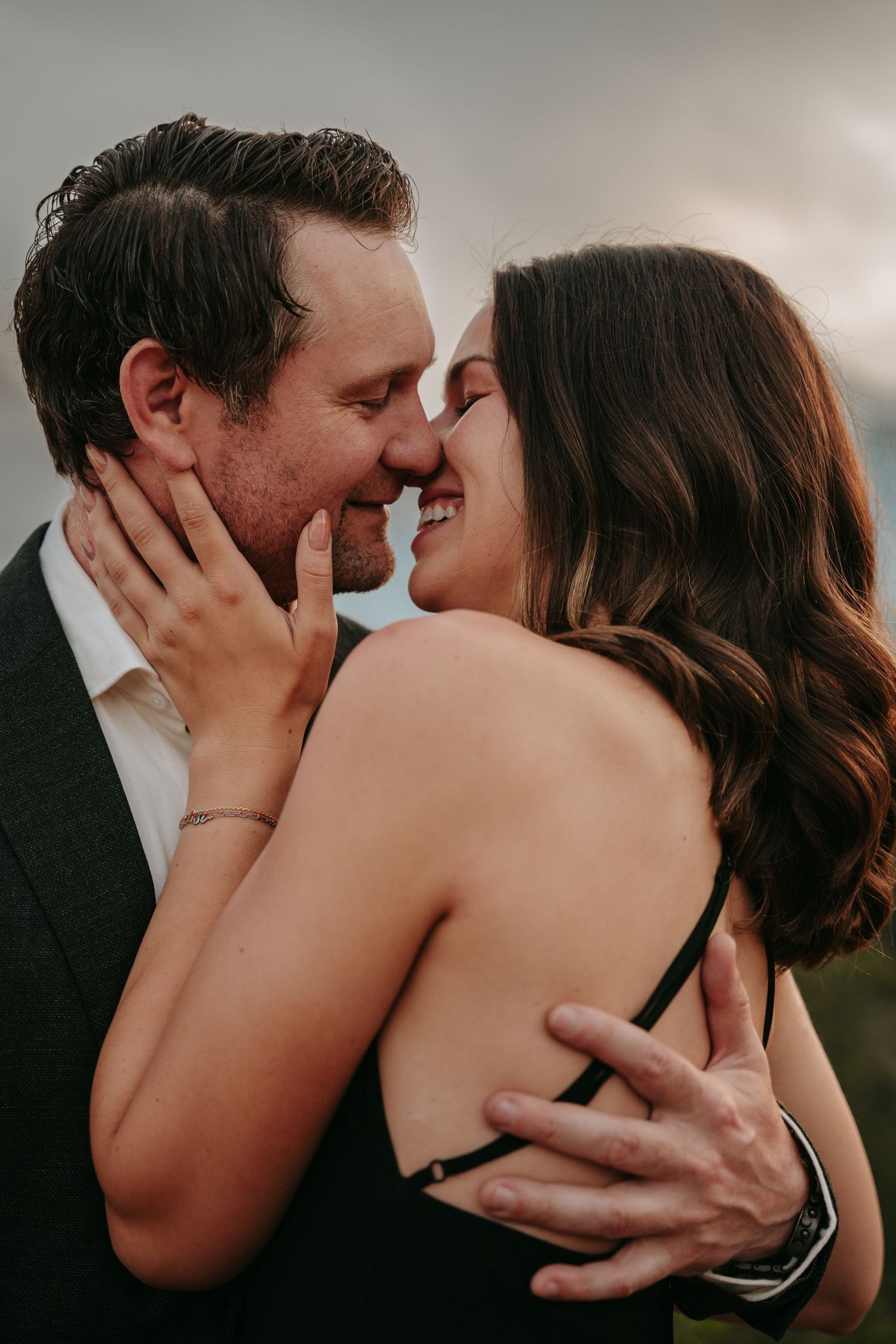 A couple shares a romantic almost-kiss embrace outdoors, the woman smiling as the man holds her close at golden hour.