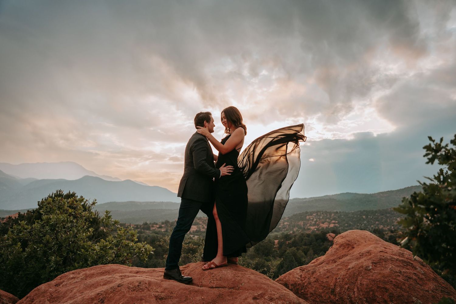 Couple embracing on red rocks at sunset with dramatic cloudy sky and mountains, woman's black dress flowing in the wind.
