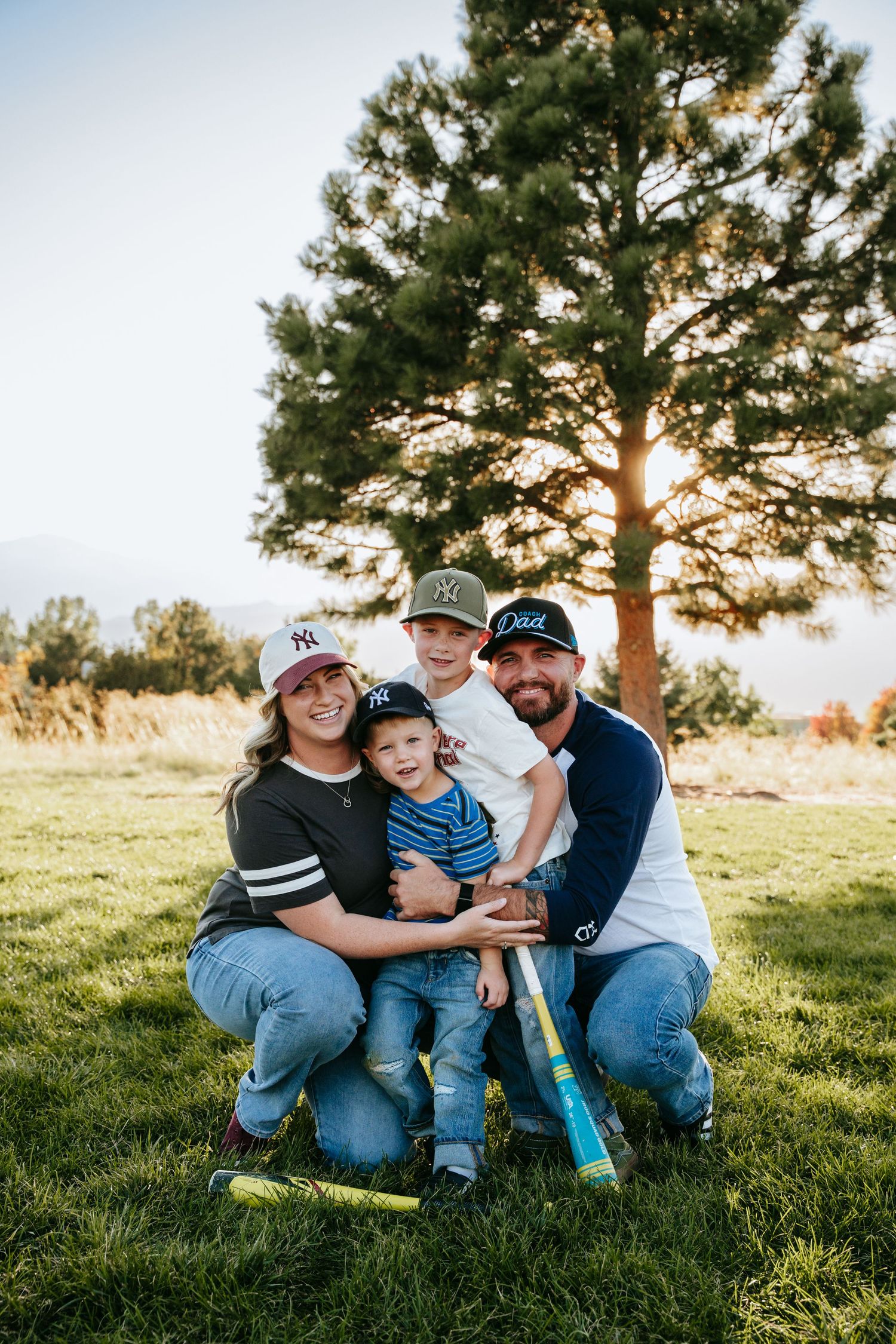 Smiling family of four in baseball caps kneel on a grassy field with bats near a tall pine tree at sunset.