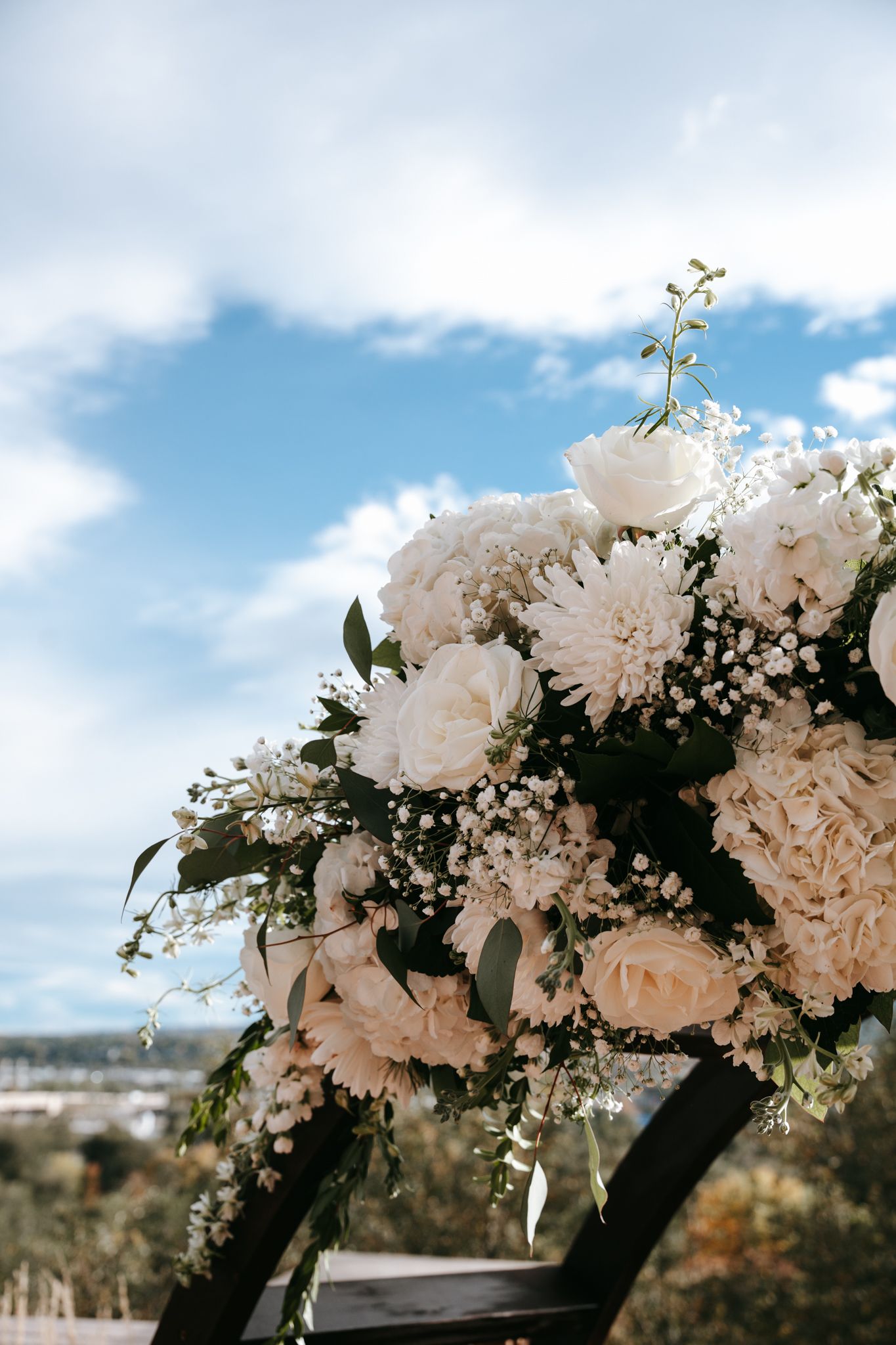 Elegant white floral wedding arch decorated with roses, dahlias, and baby's breath under blue sky.