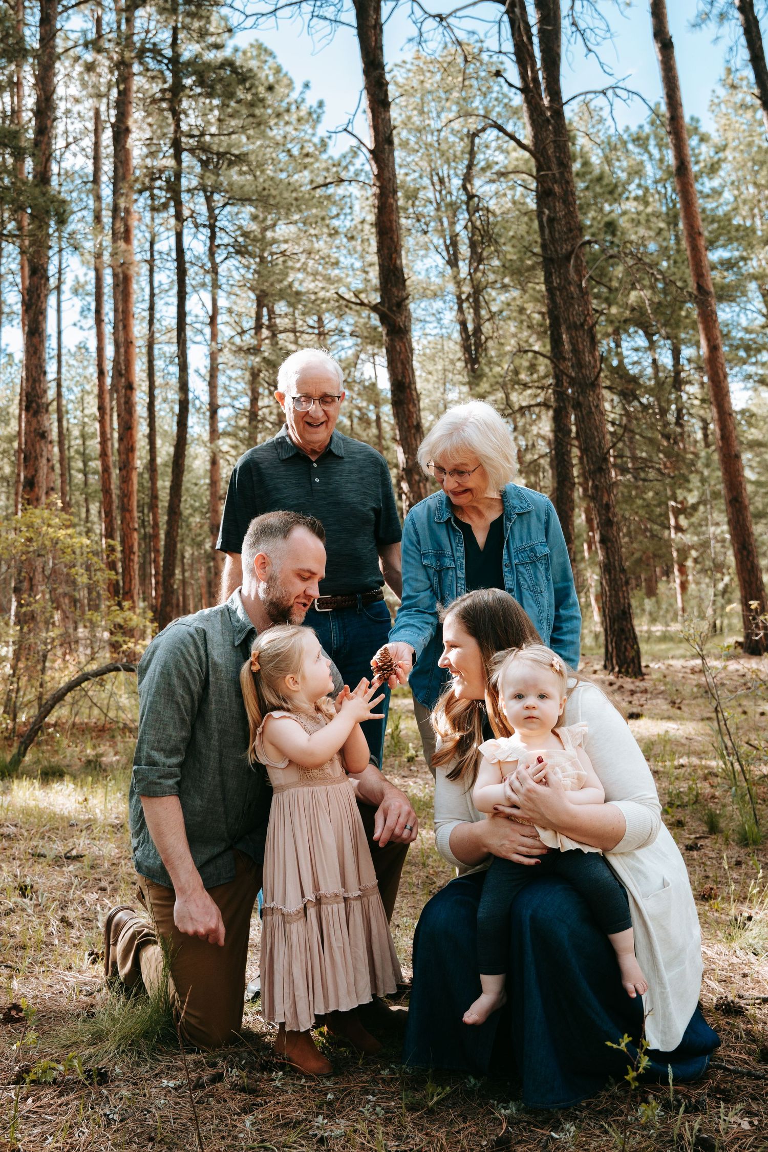 Multi-generational family smiling together in a pine forest, with grandparents, parents, and two young children.