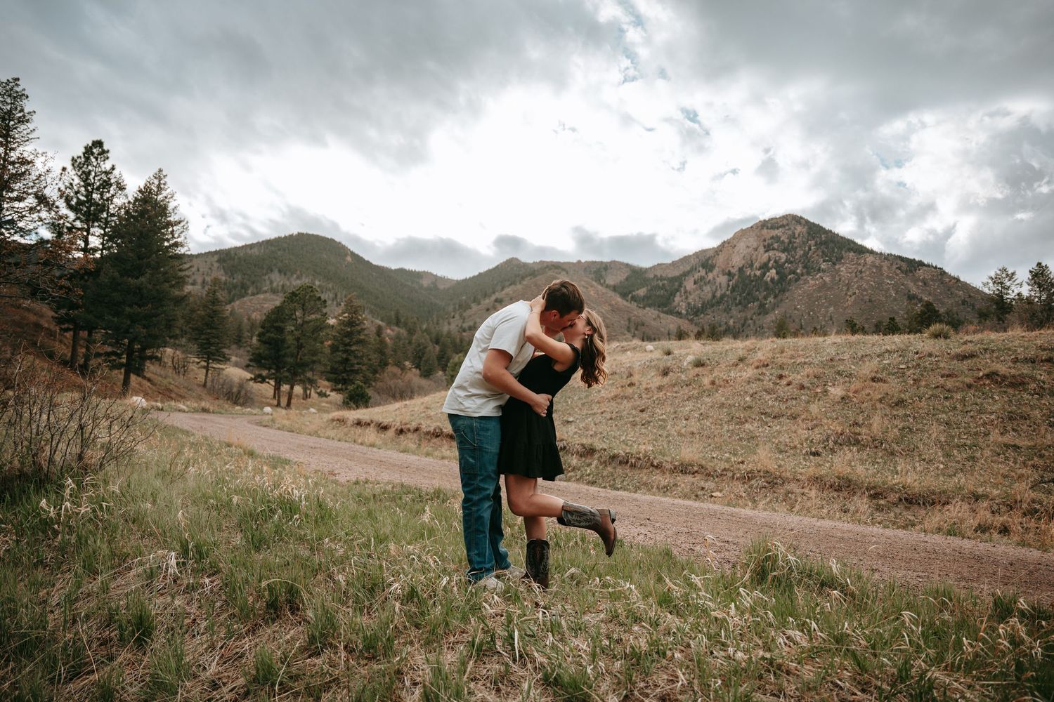 Couple sharing romantic dip kiss on dirt road with dramatic mountain landscape backdrop.