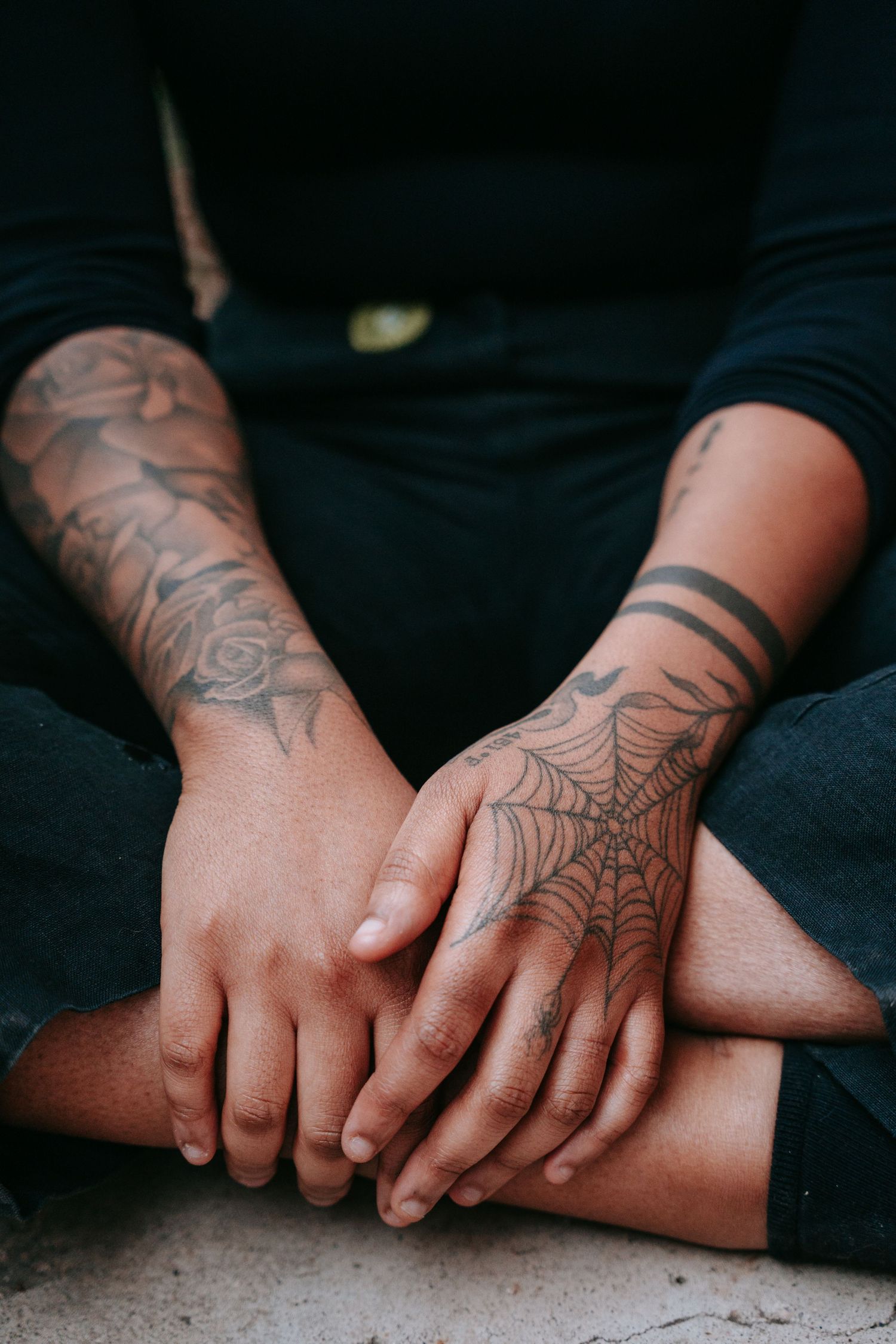 Close-up of tattooed hands resting on knees, featuring spider web and floral tattoo designs.